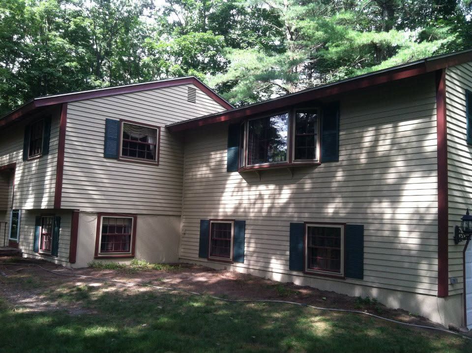 Two-story beige house with dark shutters in a wooded yard