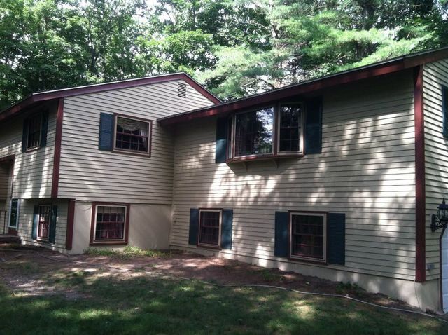 Two-story beige house with dark shutters, set among trees and grass in a wooded yard