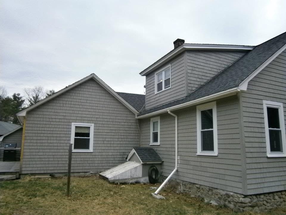 Two gray houses with a damaged white fence and debris in a grassy yard under an overcast sky