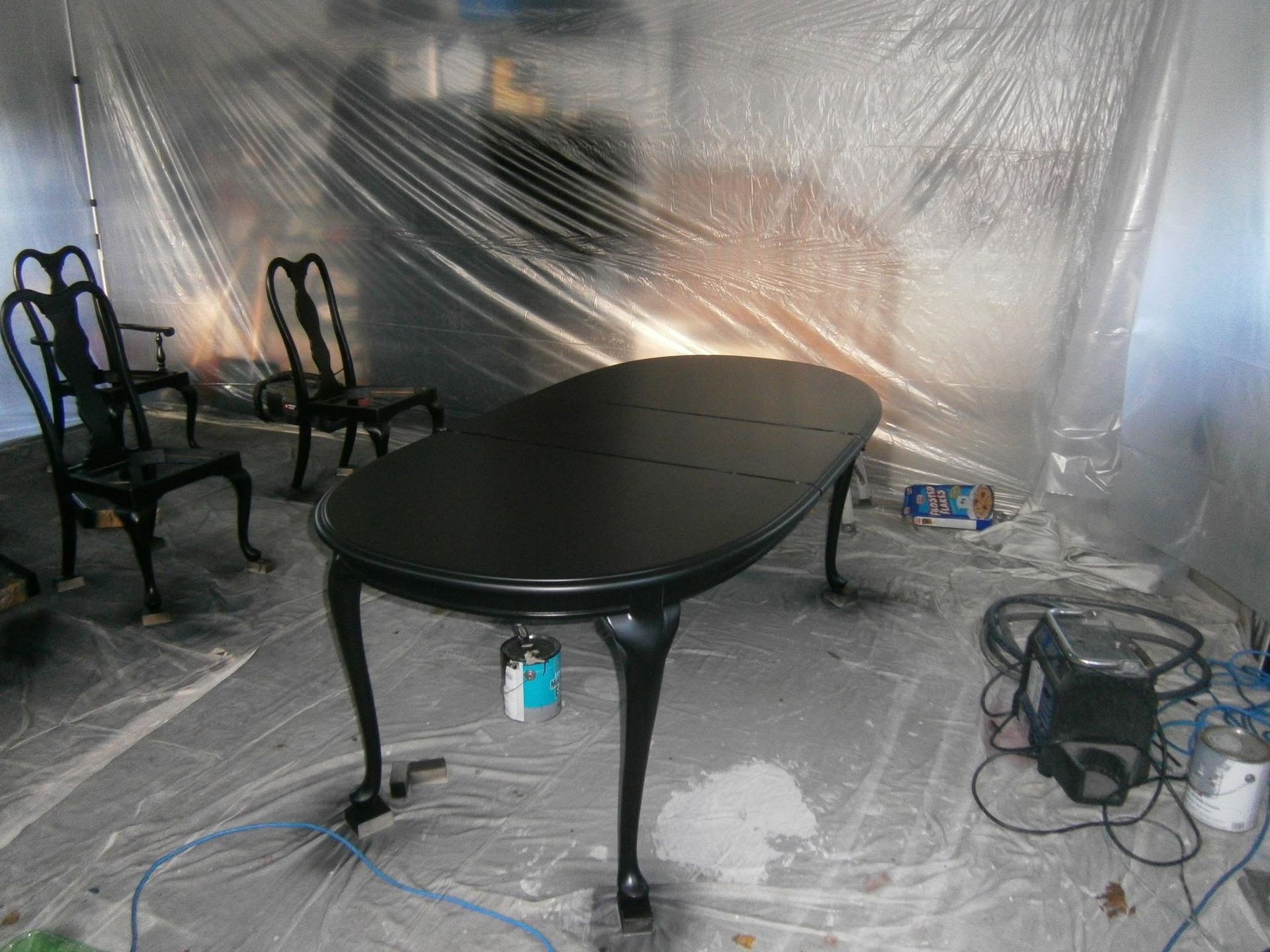 Black oval dining table and chairs in a plastic-covered room under renovation with tools on the floor