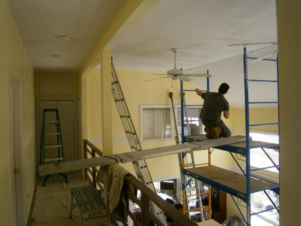 Two workers on scaffolding painting a room under renovation, with ladders and tools visible.