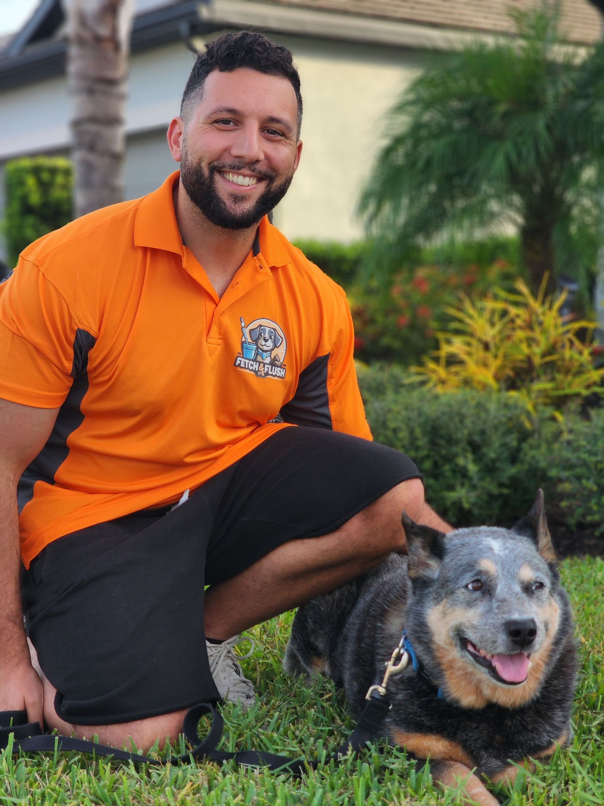 Man kneeling with a dog on grass, wearing orange polo. The dog is blue and tan with a leash. Outdoor setting.