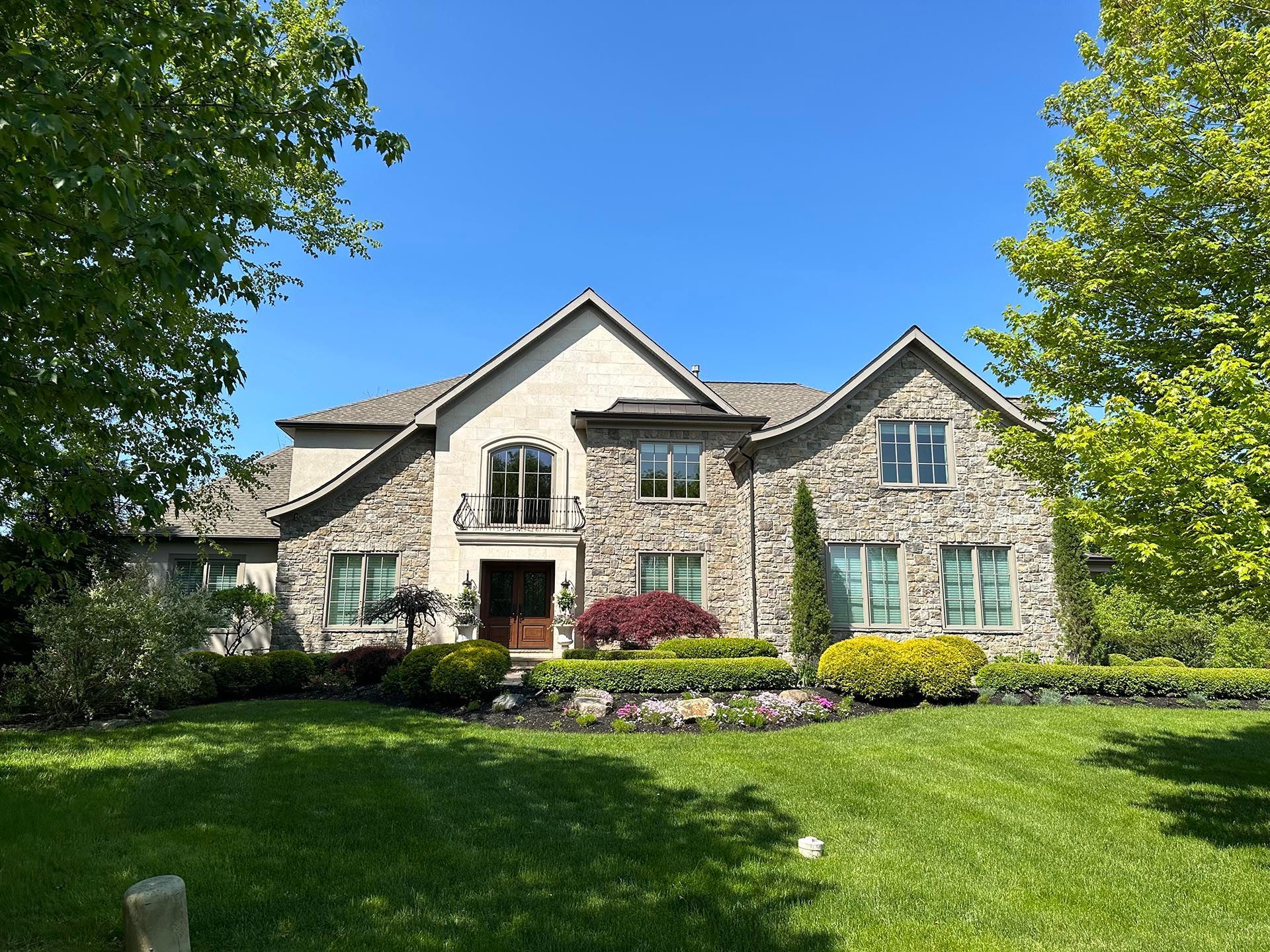 Stone house with blue shutters and a manicured lawn, framed by trees under a clear blue sky