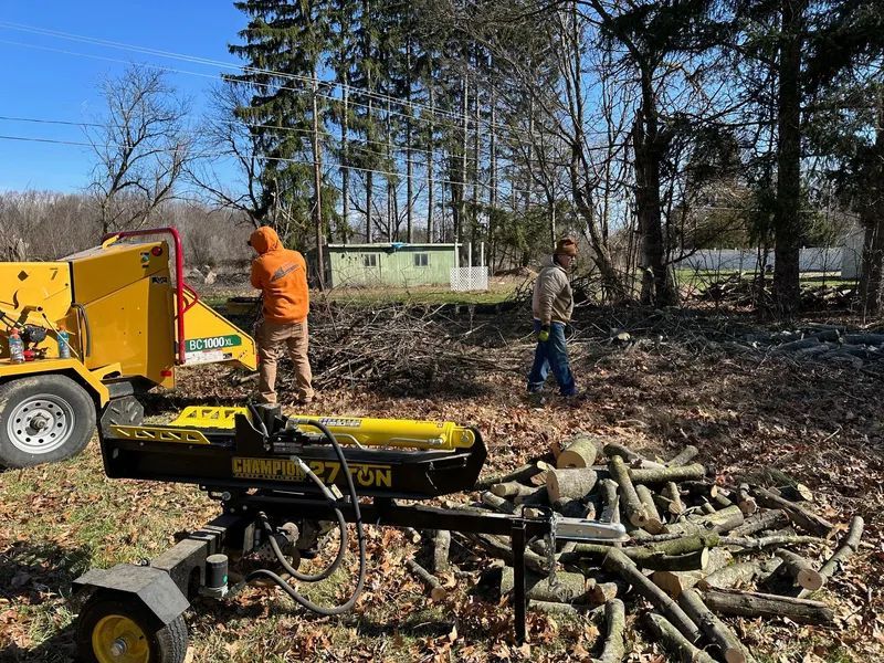 Two people working on a woodpile with a log splitter and wood chipper in a yard on a sunny day.