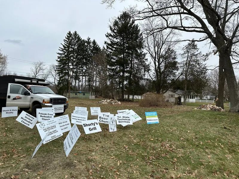 A truck and signs are in a grassy yard, with a home visible in the background. The signs appear to be protesting.
