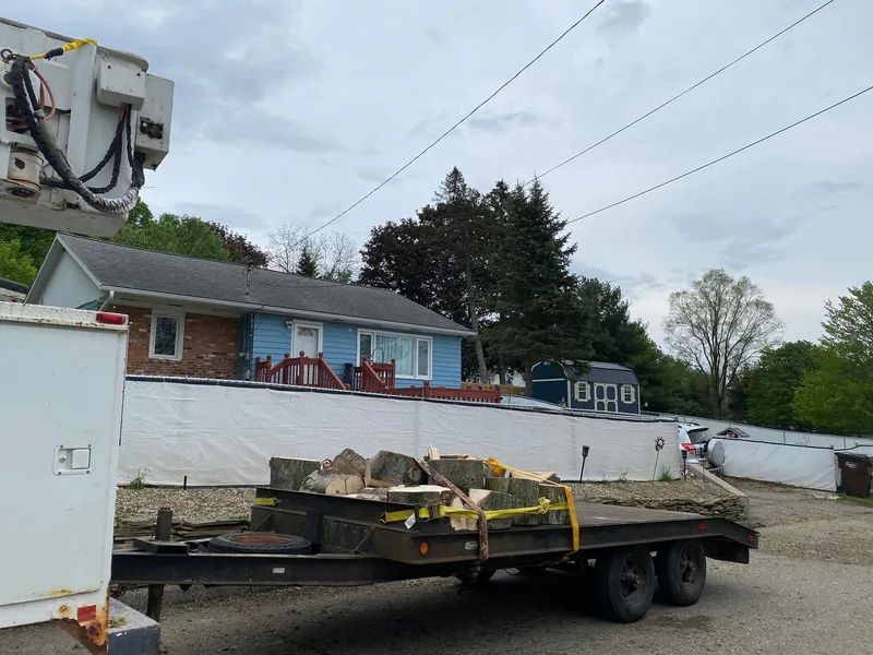 Trailer with wood, house with blue siding, power lines, and utility truck in an overcast setting.