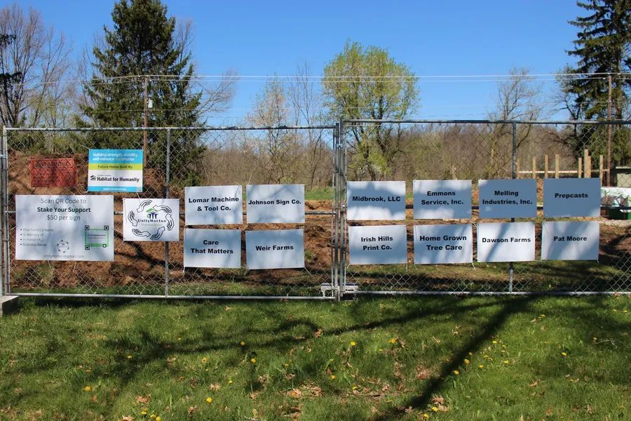 Fence with signs; sponsors listed, background of trees and grass, clear sky.