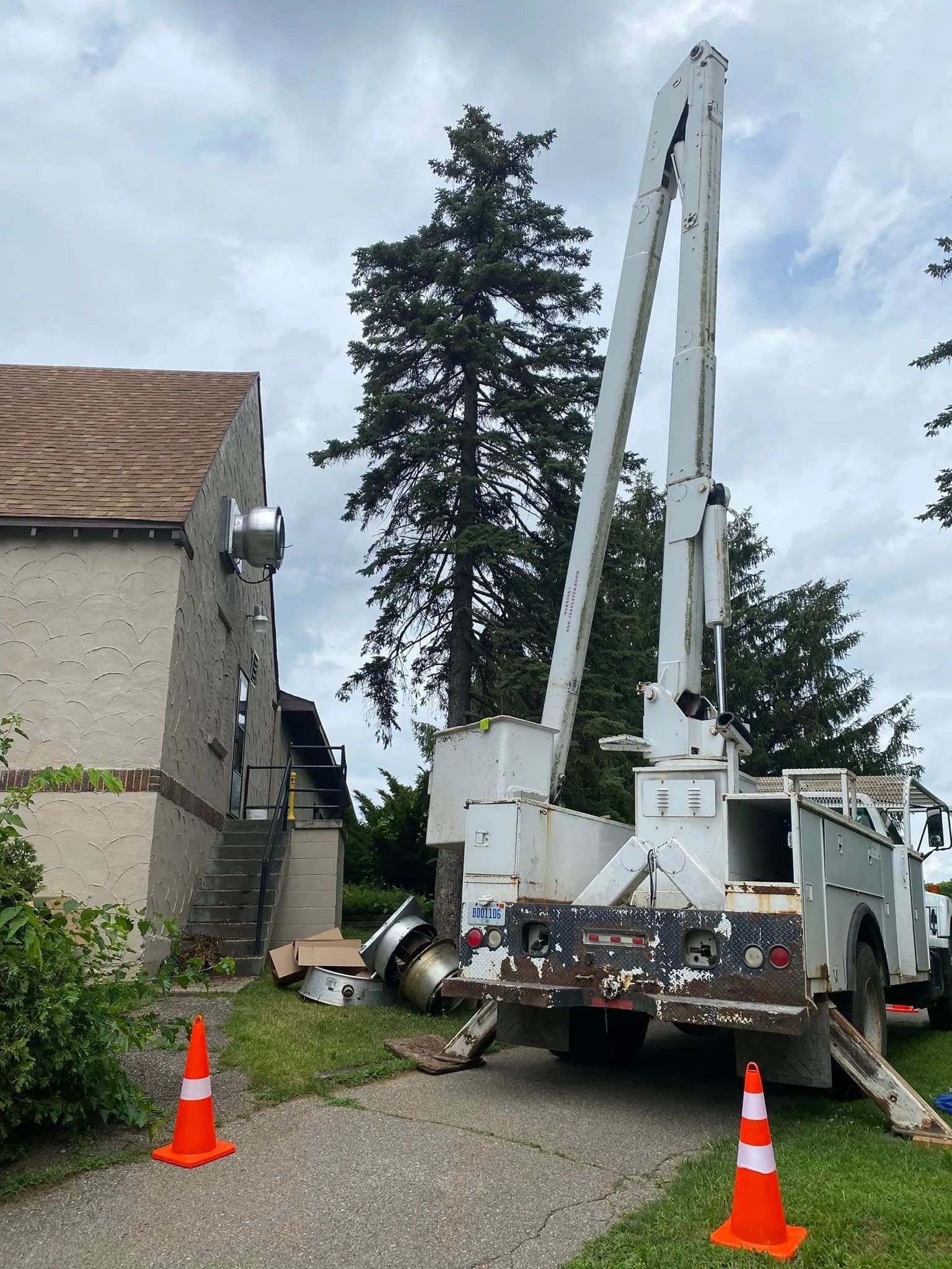 White utility truck with an extended arm near a building, next to trees. Orange traffic cones on the ground.