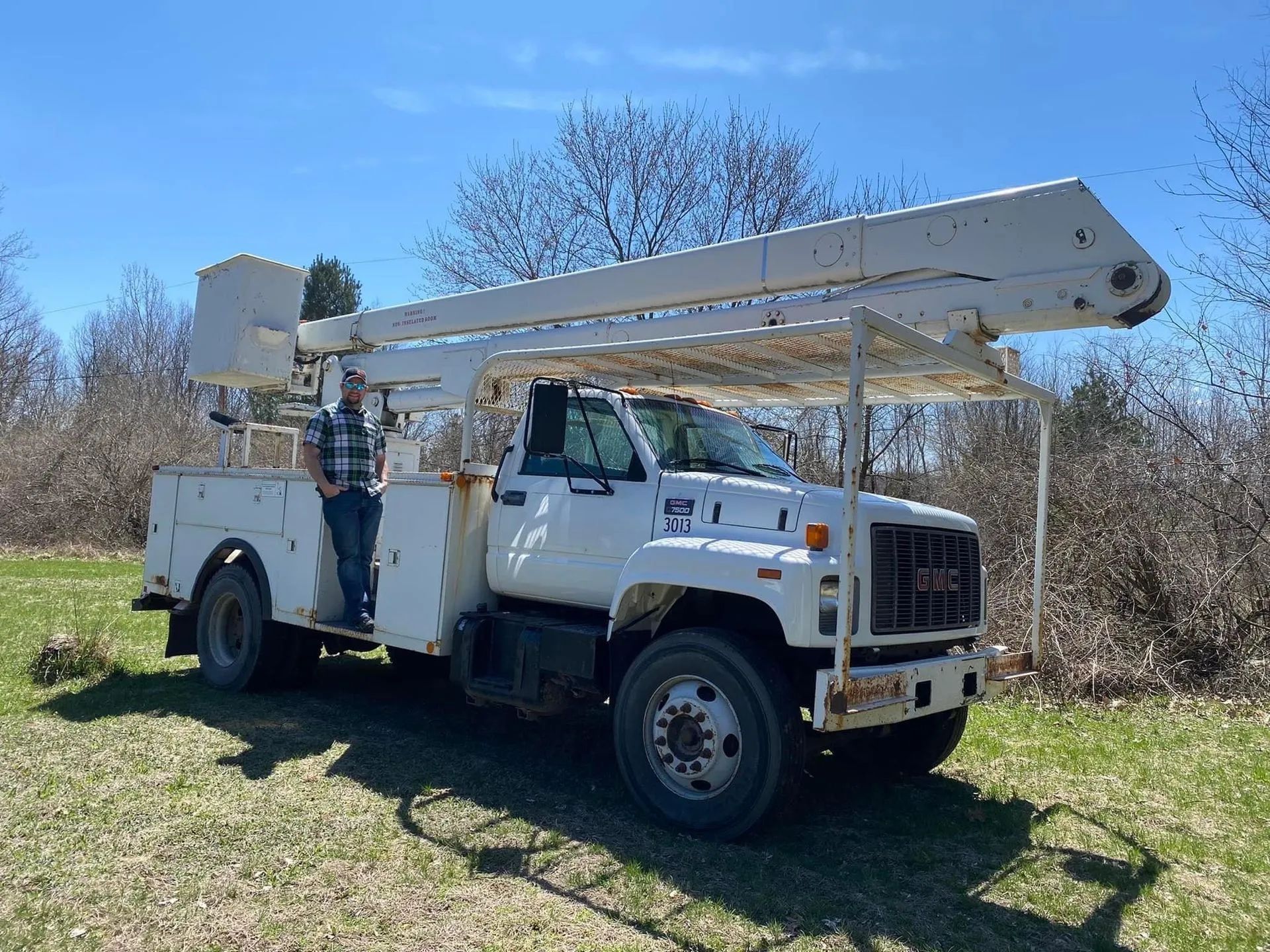 Man standing next to a white utility truck with an extended aerial lift in a grassy area on a sunny day.