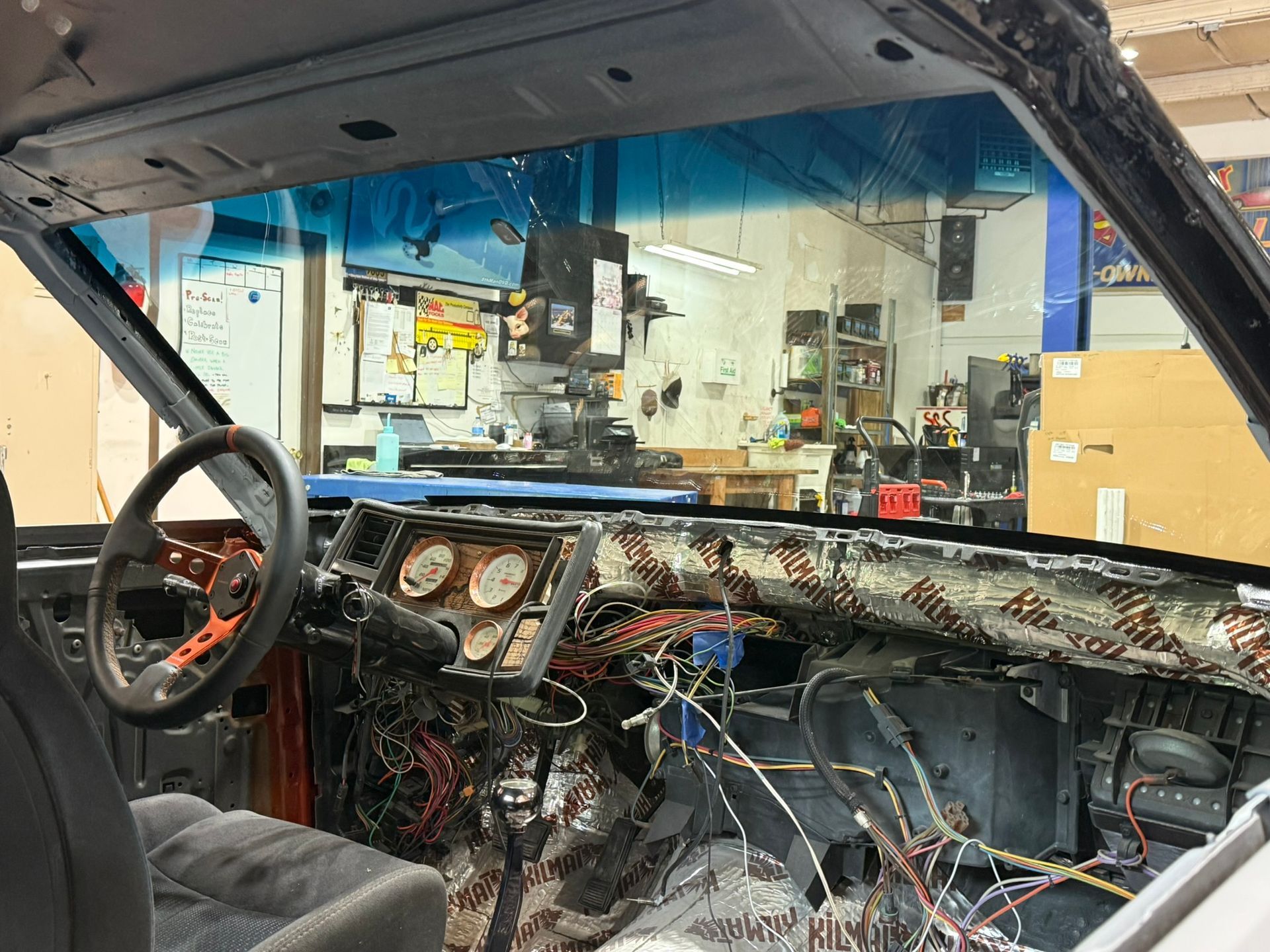 Inside a stripped car interior with exposed wiring, dashboard gauges, and a workshop visible through the window.