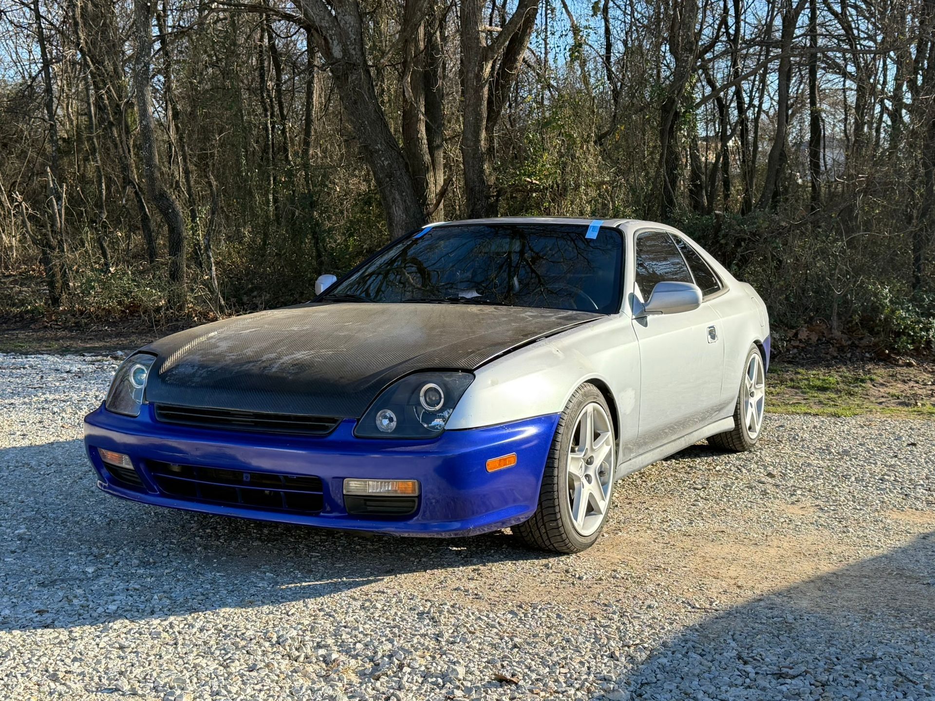 Silver and blue Honda Prelude with a black hood, parked on gravel.