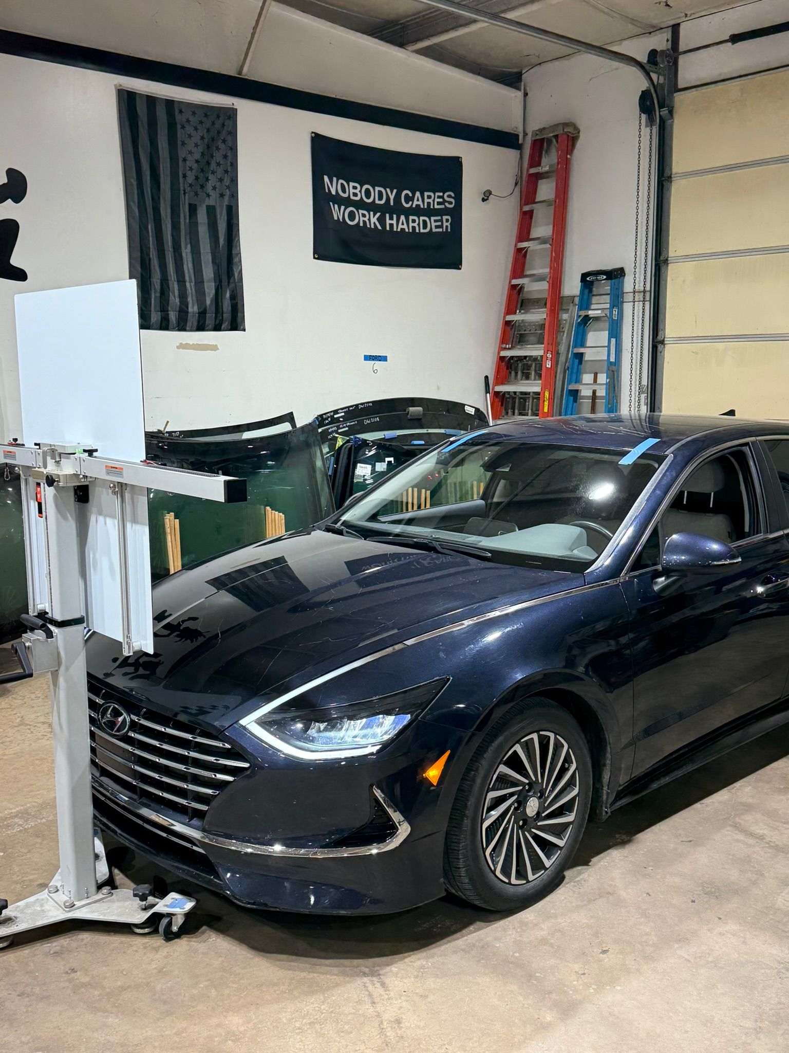 Black sedan in an auto shop bay beside a wall sign and ladder