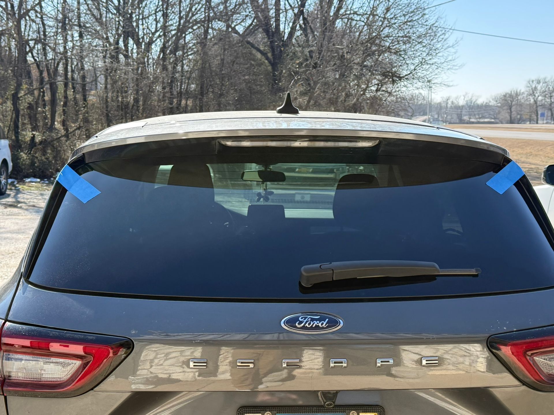 Rear view of a gray Ford Escape SUV with blue tape on the rear window in an outdoor setting.