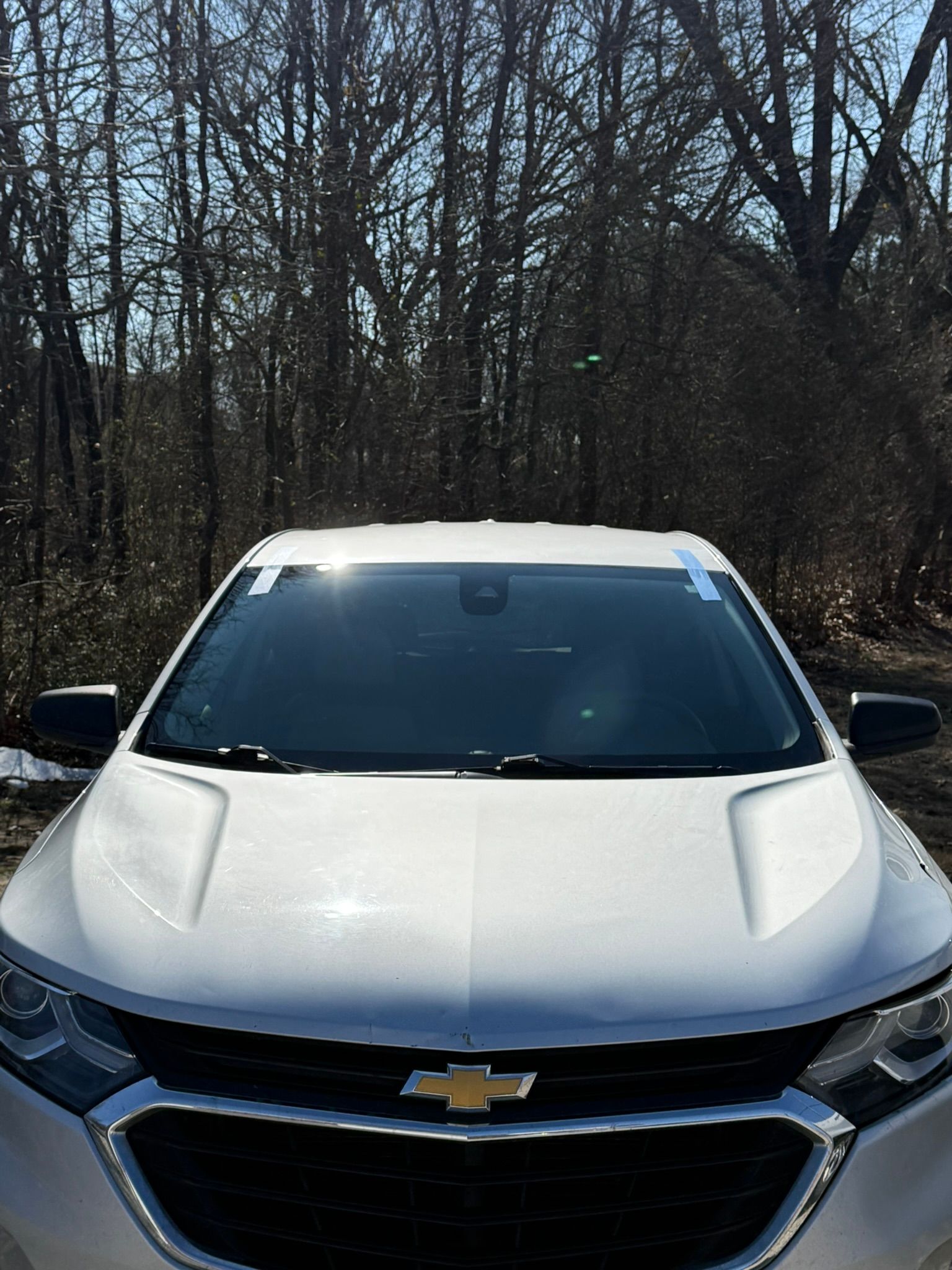 White Chevrolet SUV parked in front of a forest with a clear windshield taped.