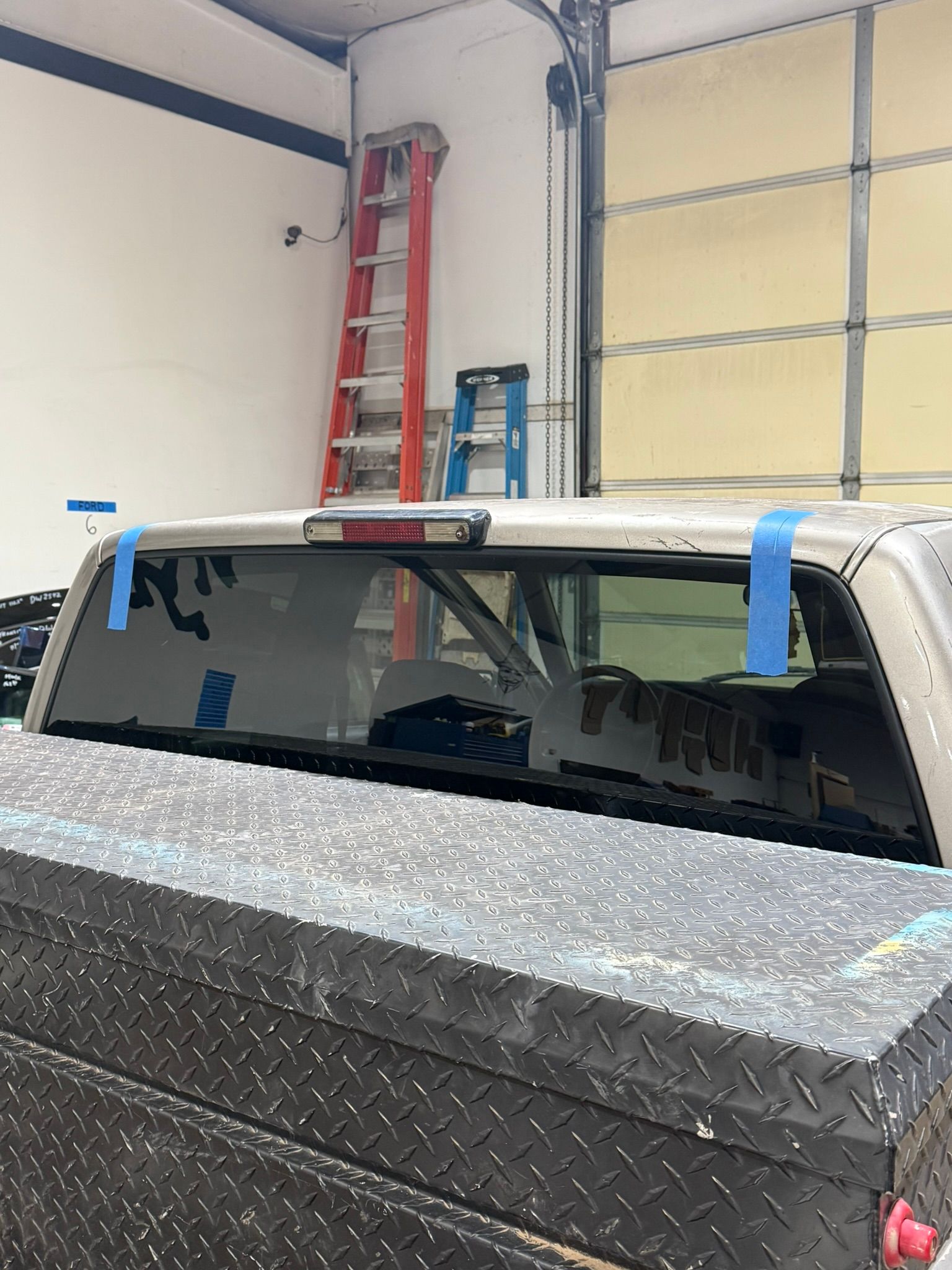 A truck's rear window with blue painter's tape on the corners, inside a garage with a red ladder in the background.
