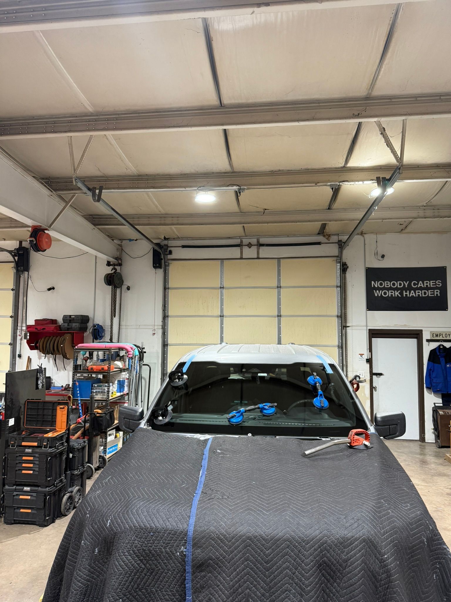 A car in a garage with its windshield covered by a protective blanket and suction tools attached for replacement.