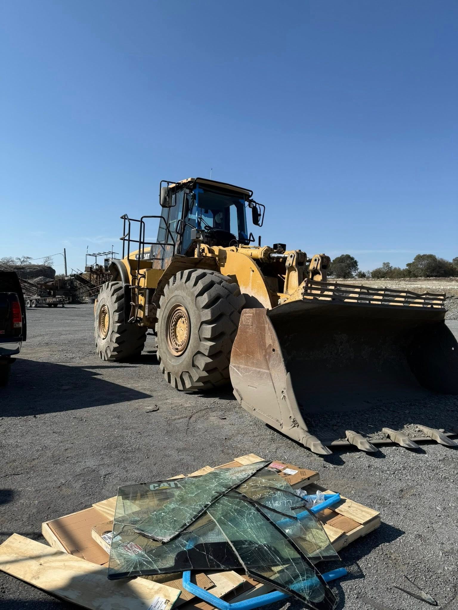 Yellow loader with raised bucket on gravel, blue sky in background. Debris and broken glass in front.