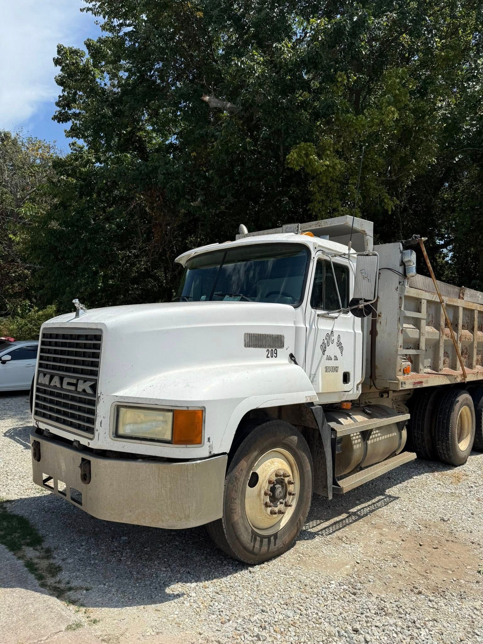 White Mack dump truck parked on gravel in front of trees, cloudy sky.