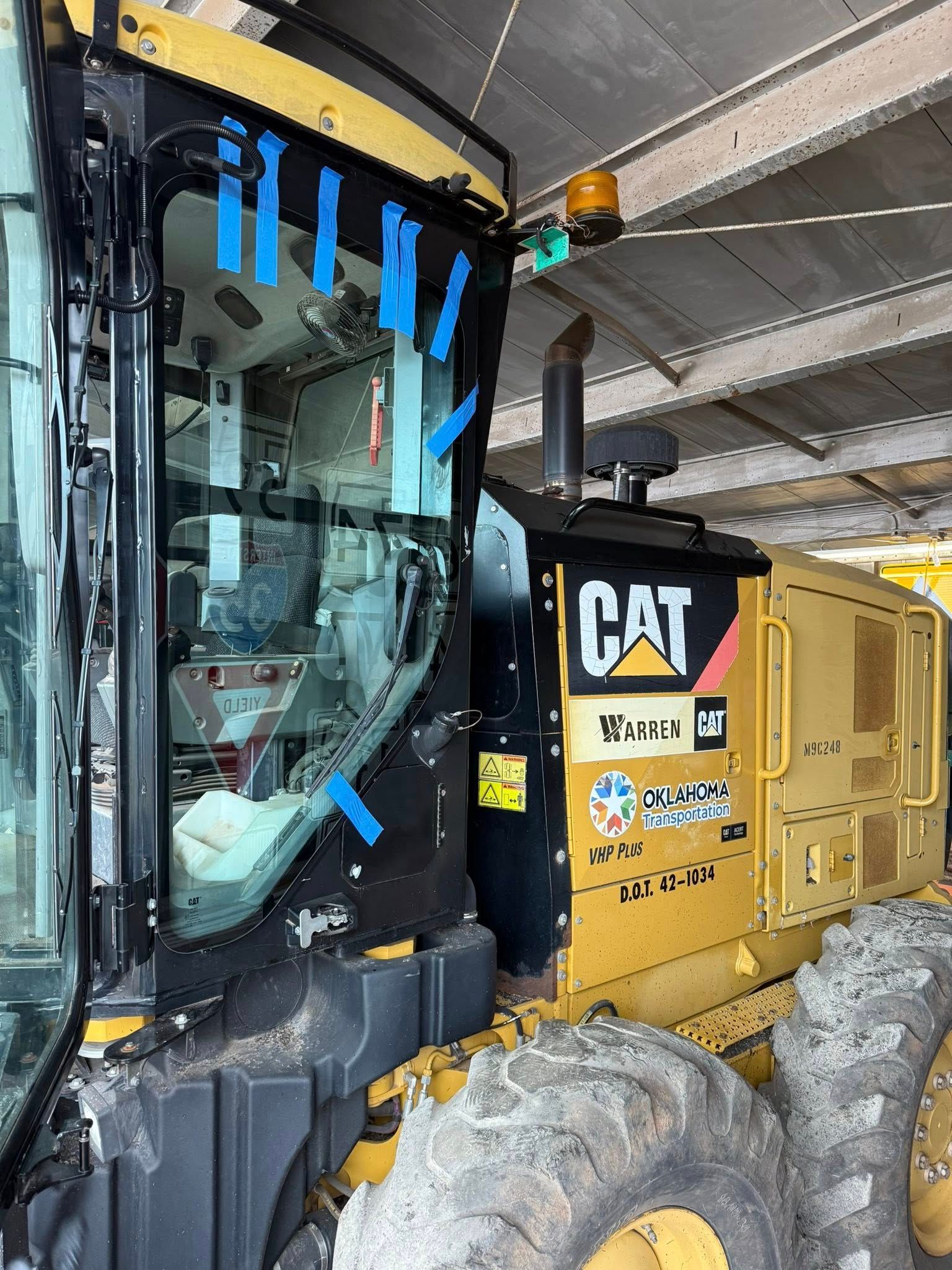 Yellow Caterpillar motor grader with open cab door, taped glass, in a sheltered area.
