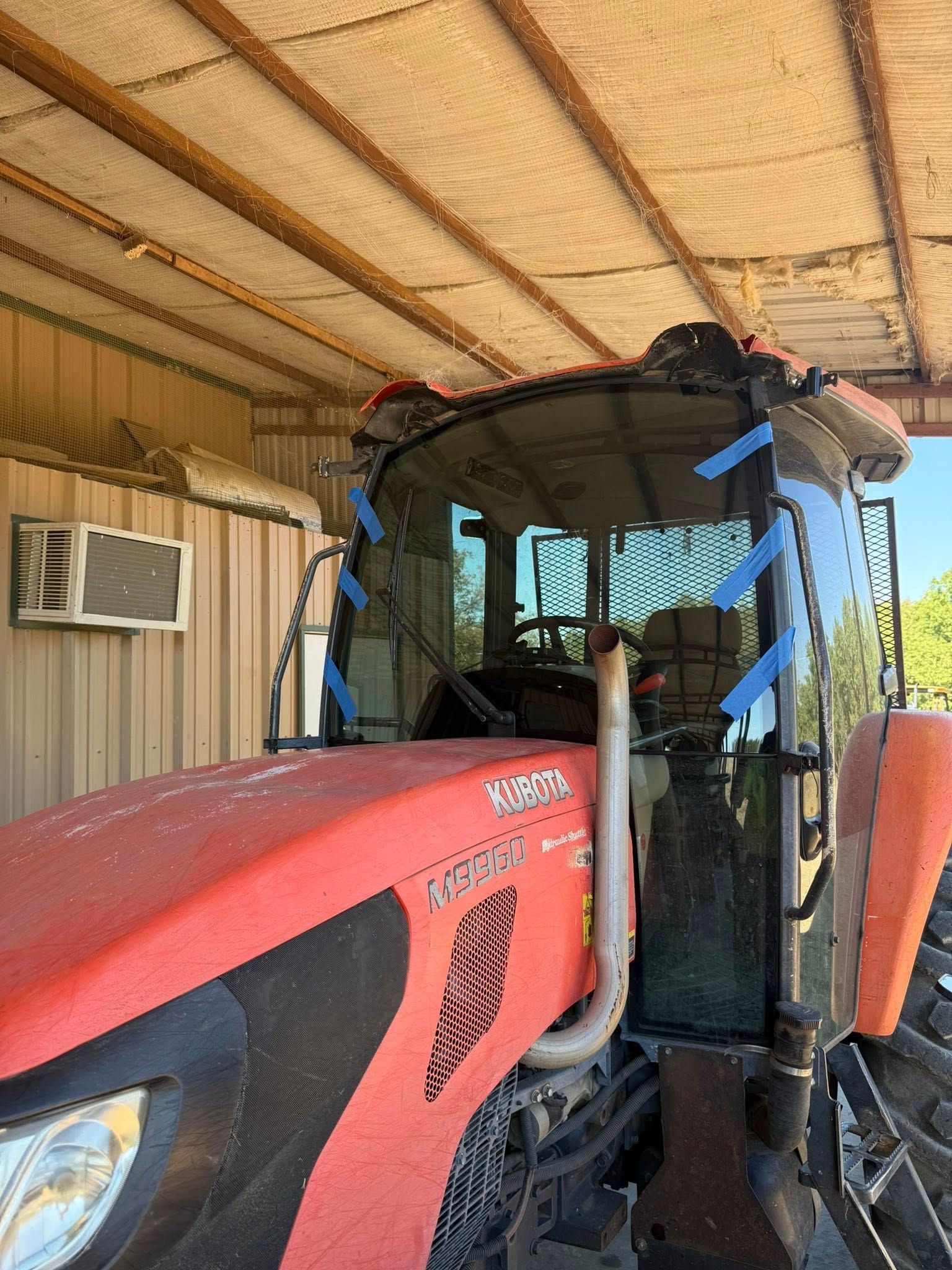 Red Kubota tractor with a glass cab, under a shed roof. Blue tape on the glass.