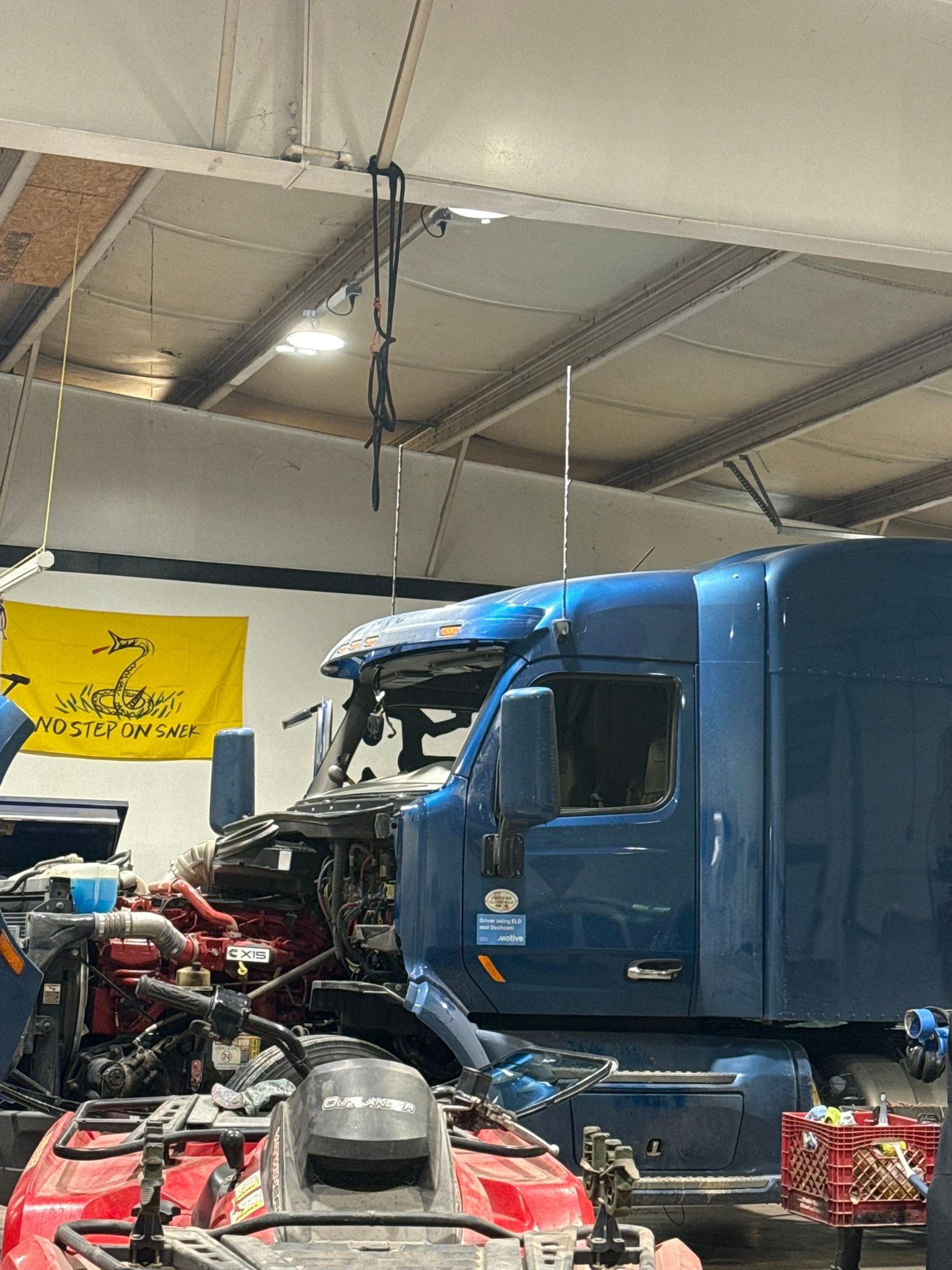 Blue semi-truck cab in a repair shop. Machinery and tools visible, yellow flag on wall.