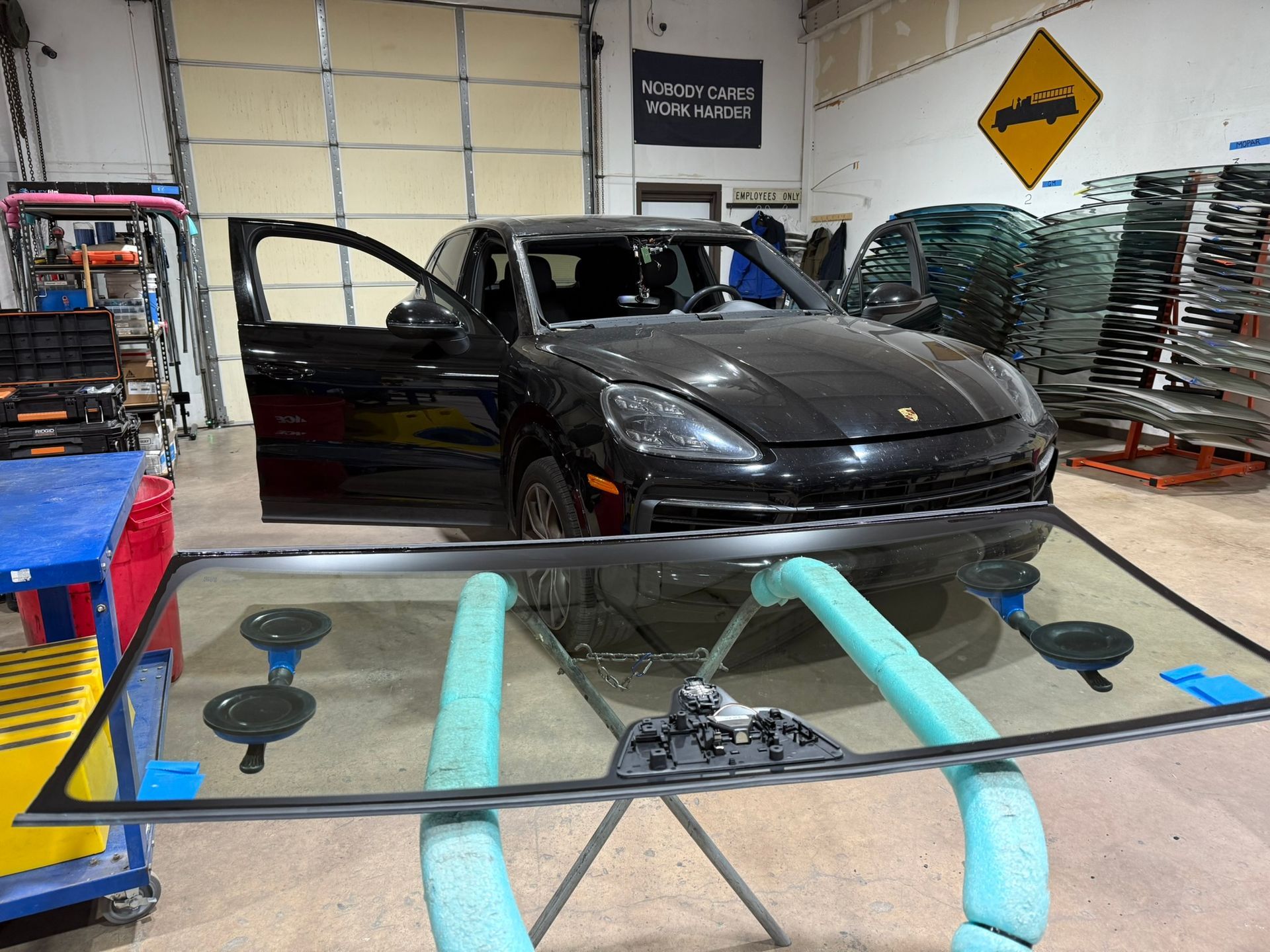 A black Porsche SUV with an open door, next to a windshield on a stand in a repair shop.
