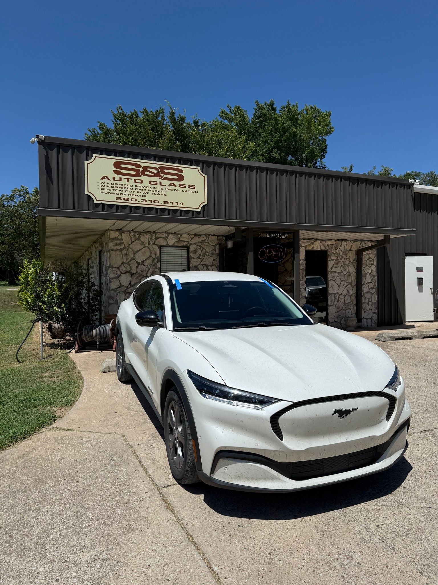 White Ford Mustang Mach-E electric vehicle parked in front of an auto repair shop on a sunny day.