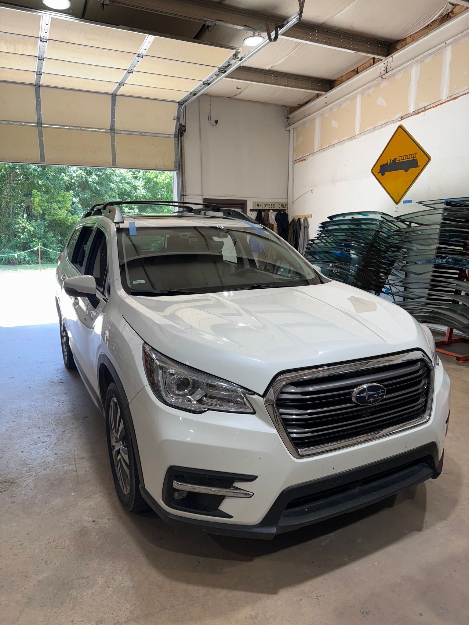 White Subaru SUV parked inside a workshop, with a caution sign on the wall, glass panels in the background.