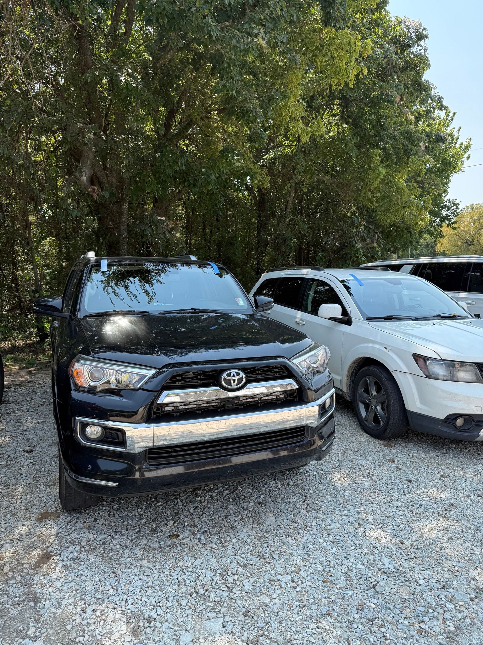 Black Toyota 4Runner and white Dodge Journey parked on gravel under trees.