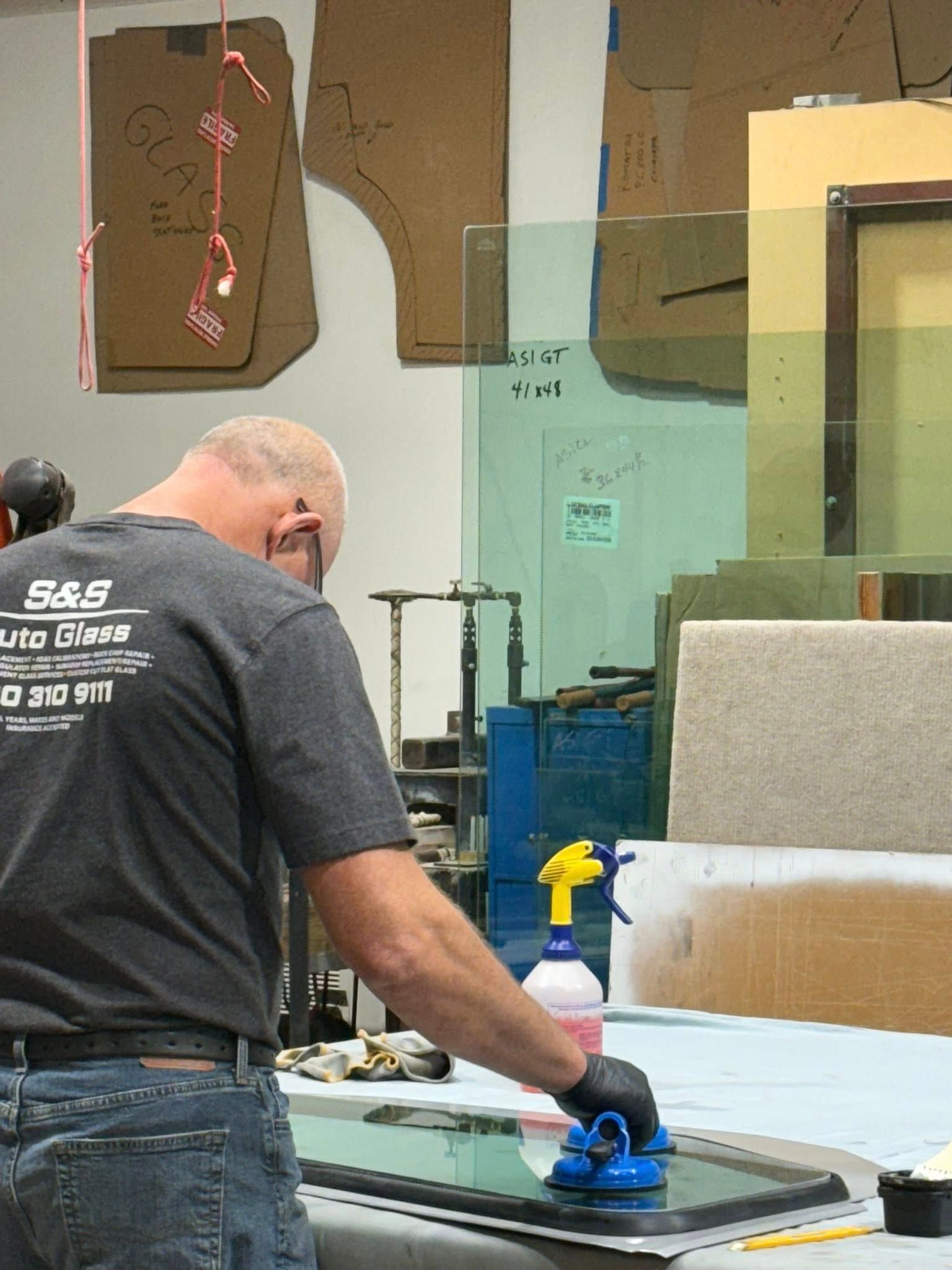 Man in a gray shirt cleaning a car window. Spray bottle and tools on a table. Workshop setting.