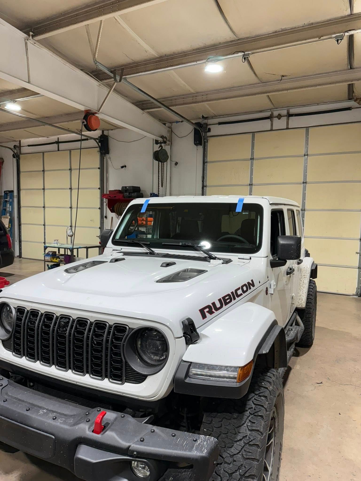White Jeep Rubicon in a garage; blue tape marks windshield.