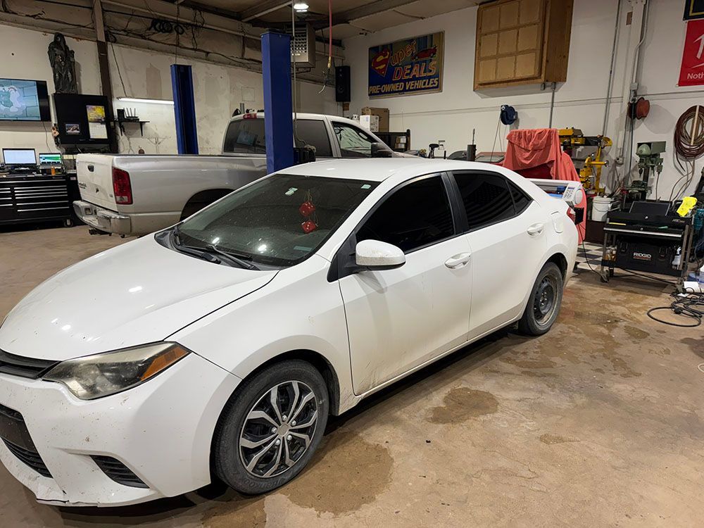 White sedan in an auto shop garage, with a pickup truck and tools in the background