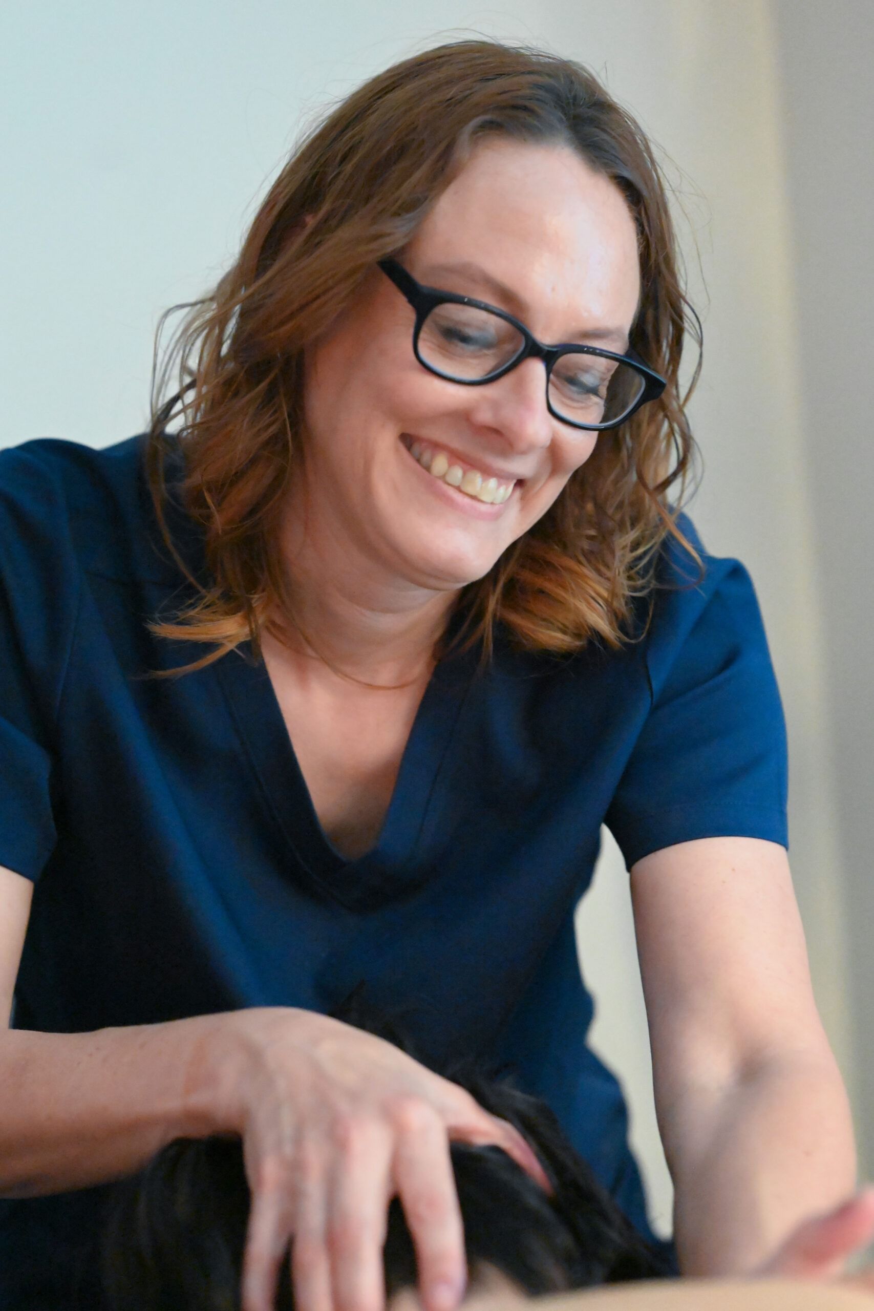 Woman in blue scrubs smiles while working on a patient, wearing glasses.