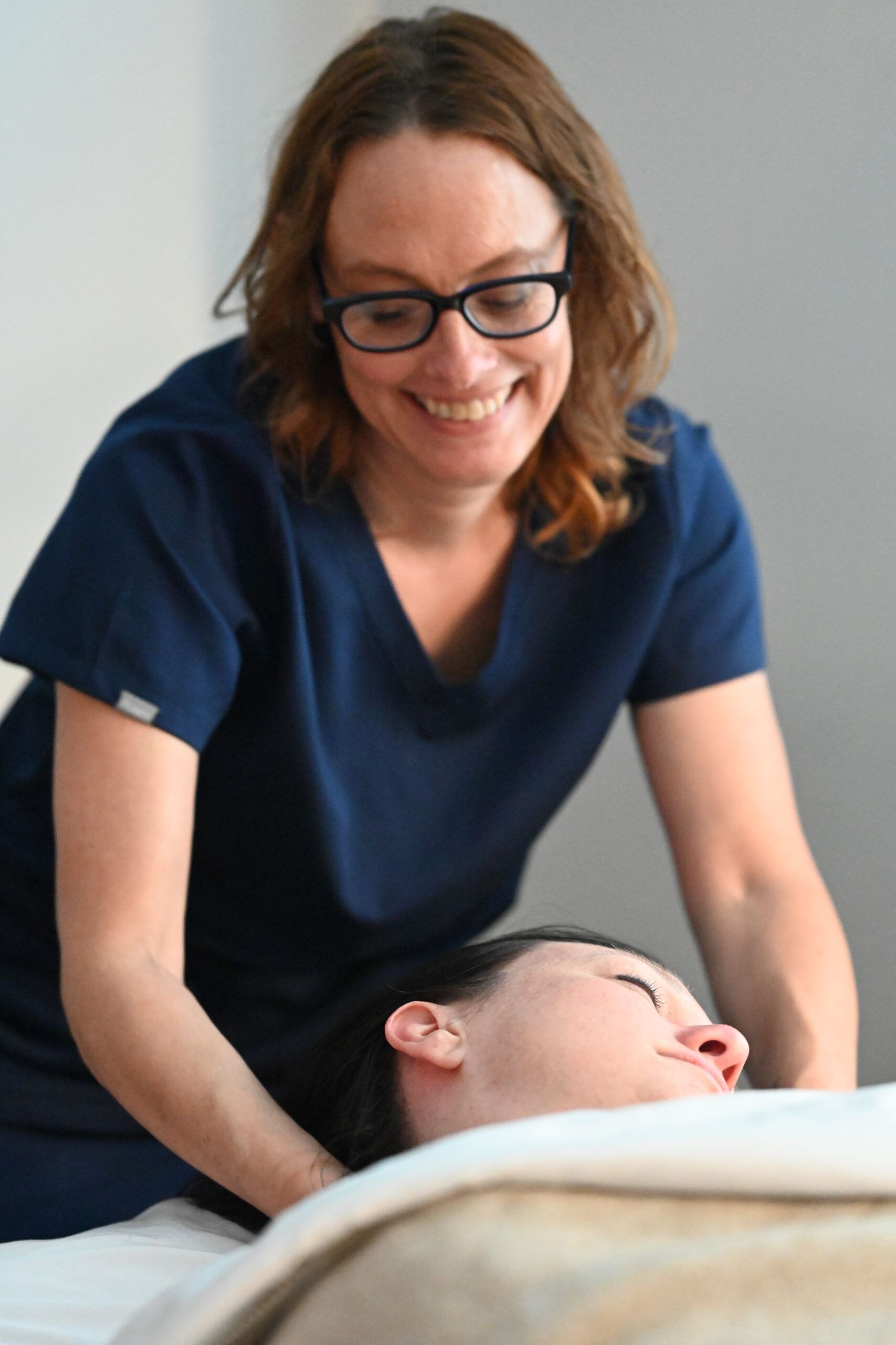 Woman in blue scrubs smiles while providing treatment to a person lying on a massage table.