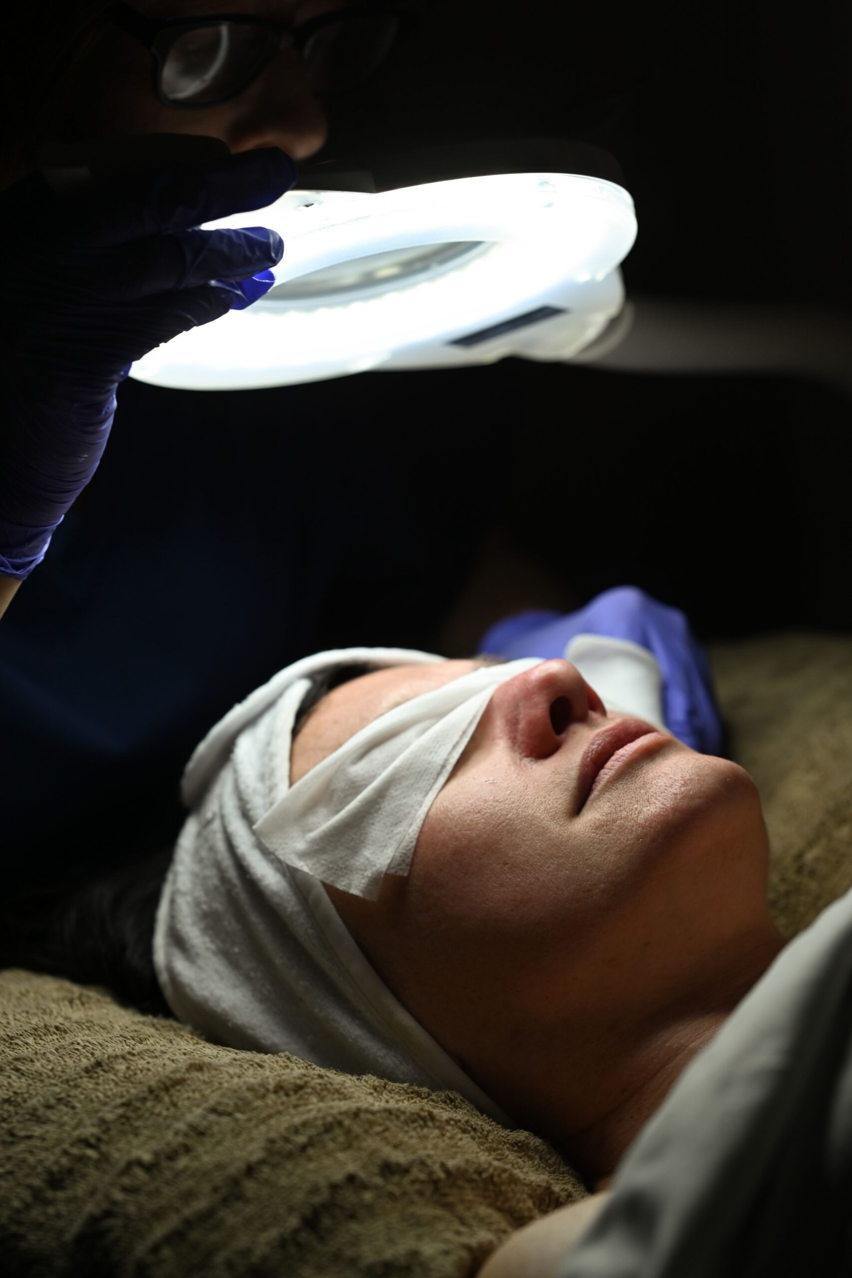 Person with eyes covered by gauze, under bright ring light. Skin care treatment.