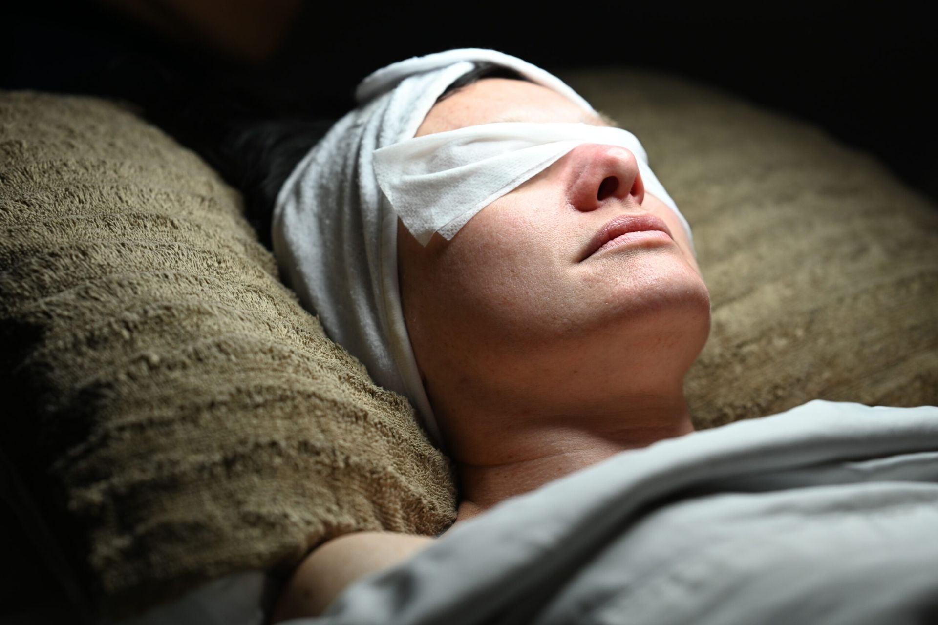 Woman with bandage over eyes, lying on a pillow. Wrapped in white towel, looking relaxed in dimly lit setting.