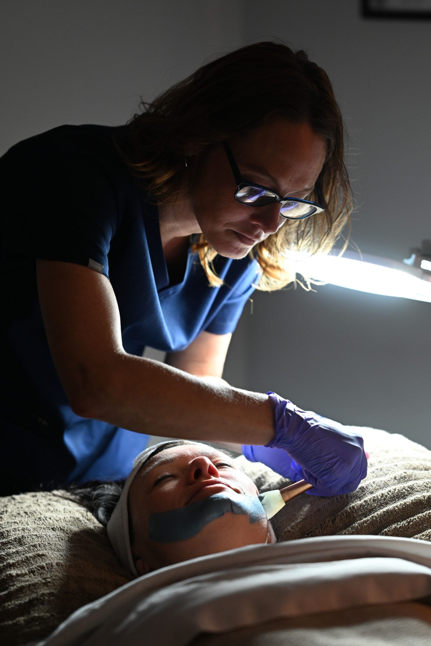 Woman in scrubs performing facial treatment on a client lying down.