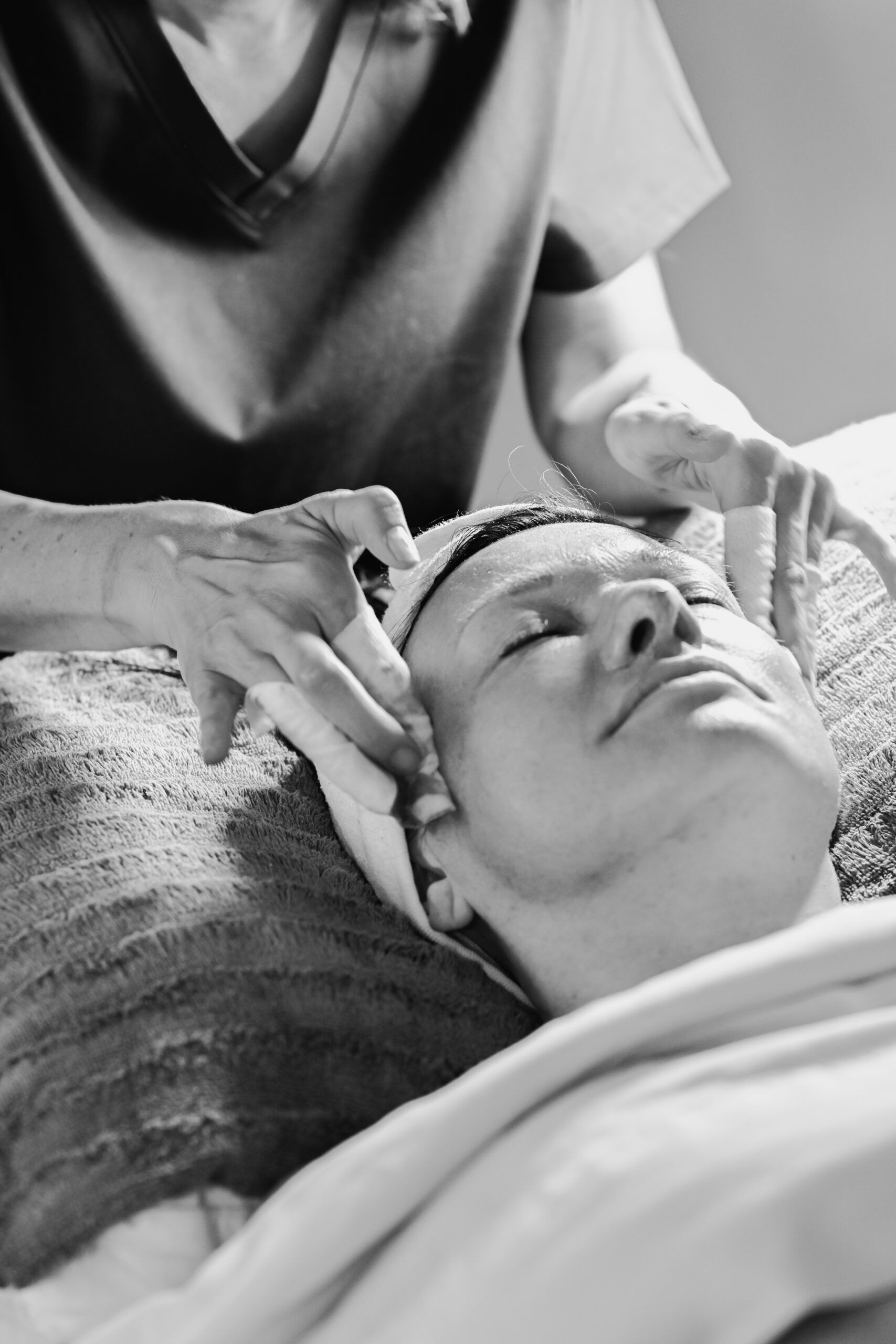 Person receiving facial treatment, head wrapped, eyes closed, hands on face by a spa employee.