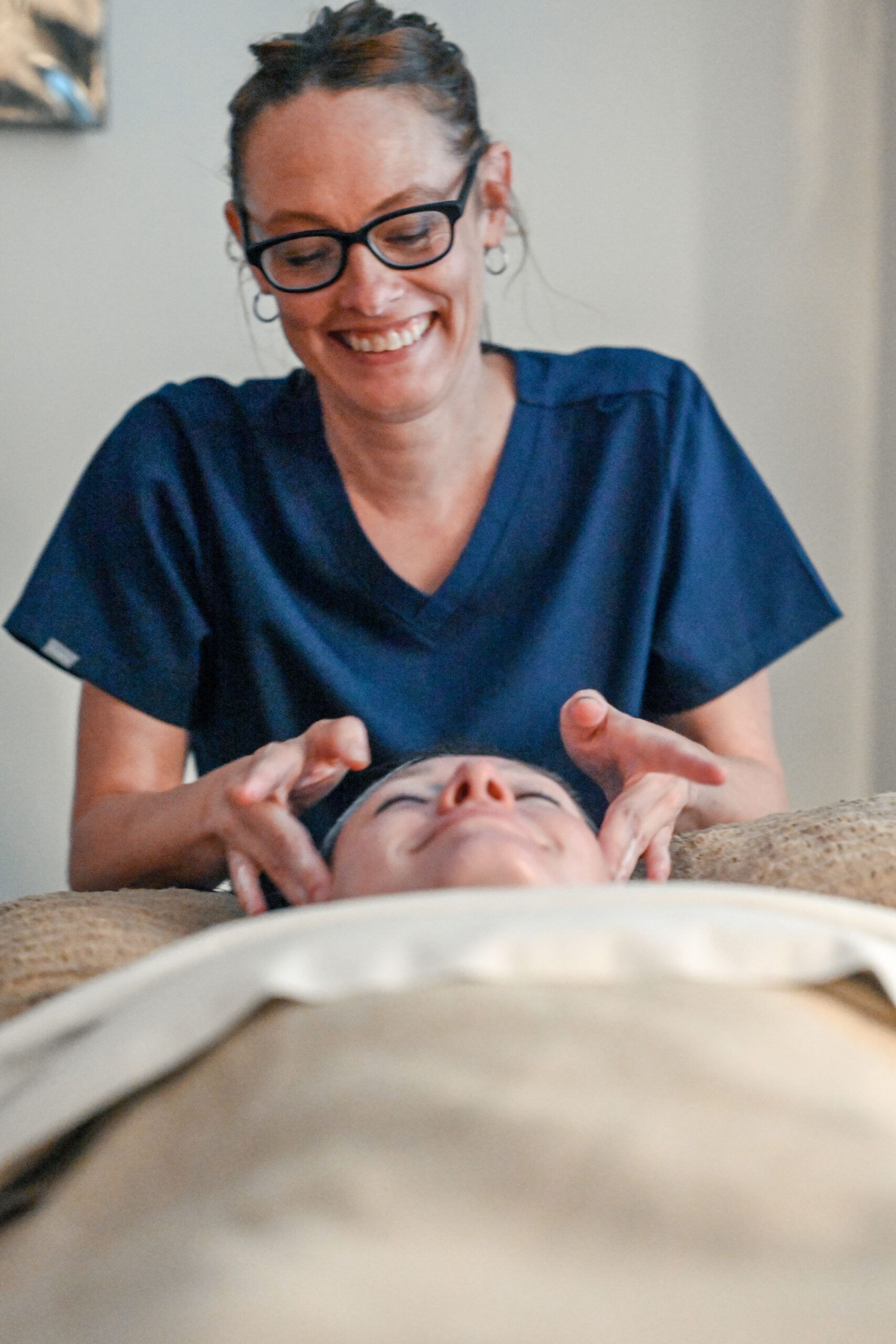 Woman in scrubs smiling, providing a facial massage to a client lying down.