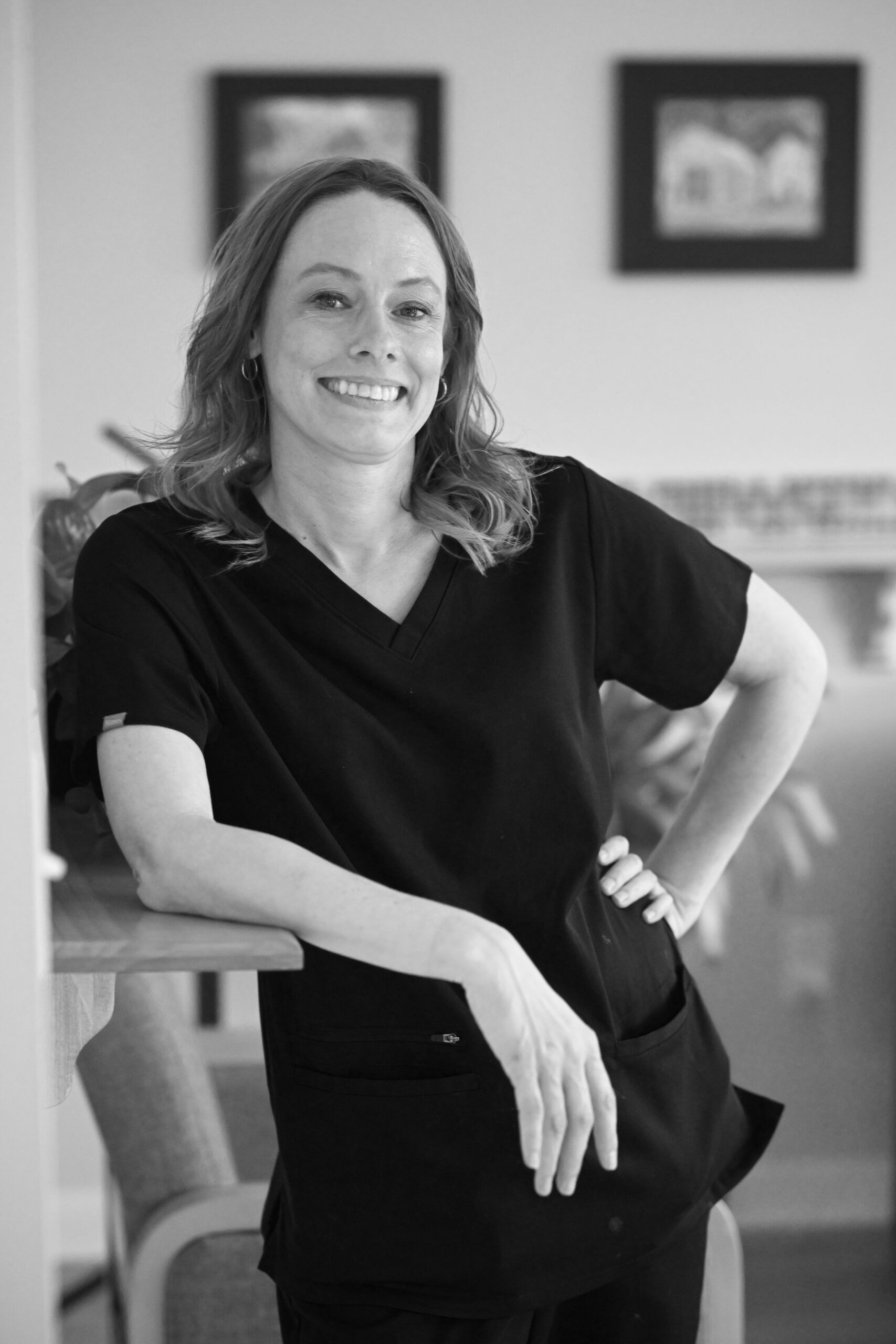 Woman in black scrubs smiles, leaning on a table.  She stands indoors with artwork in the background.