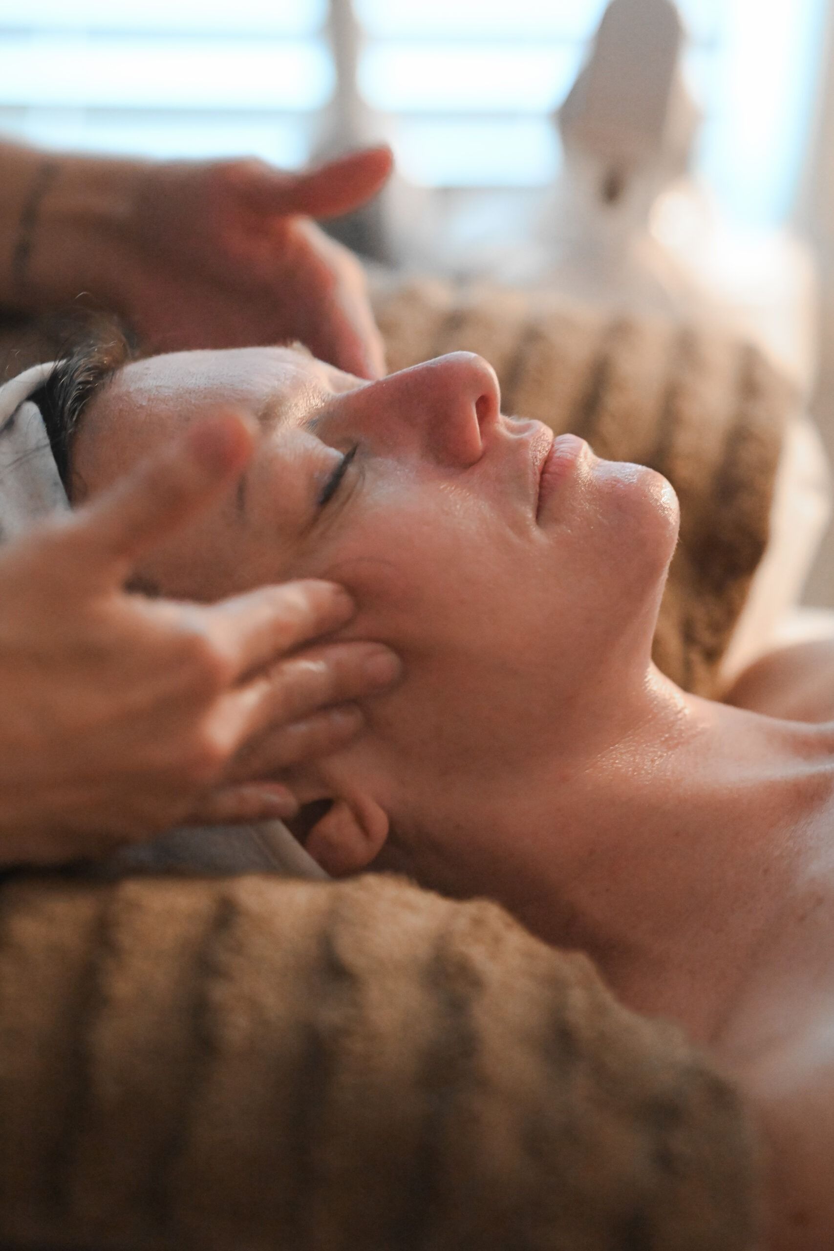 Person receiving facial massage. Face is relaxed, eyes closed. Hands massaging temples and cheek.