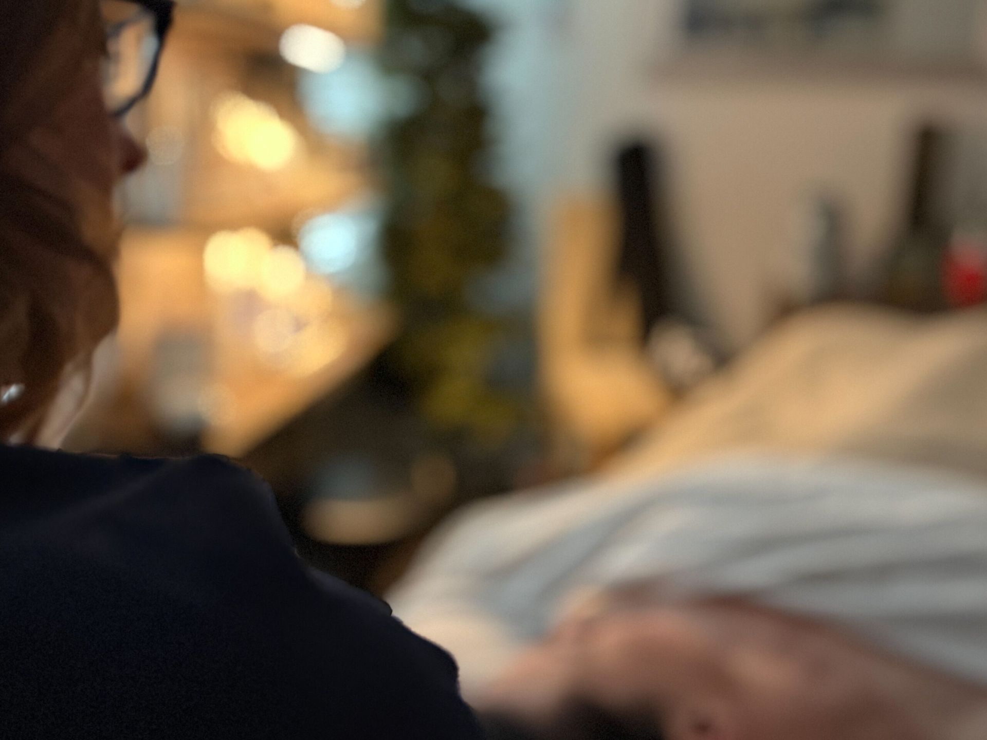 Woman with glasses looking at a person lying down indoors, possibly giving a massage.