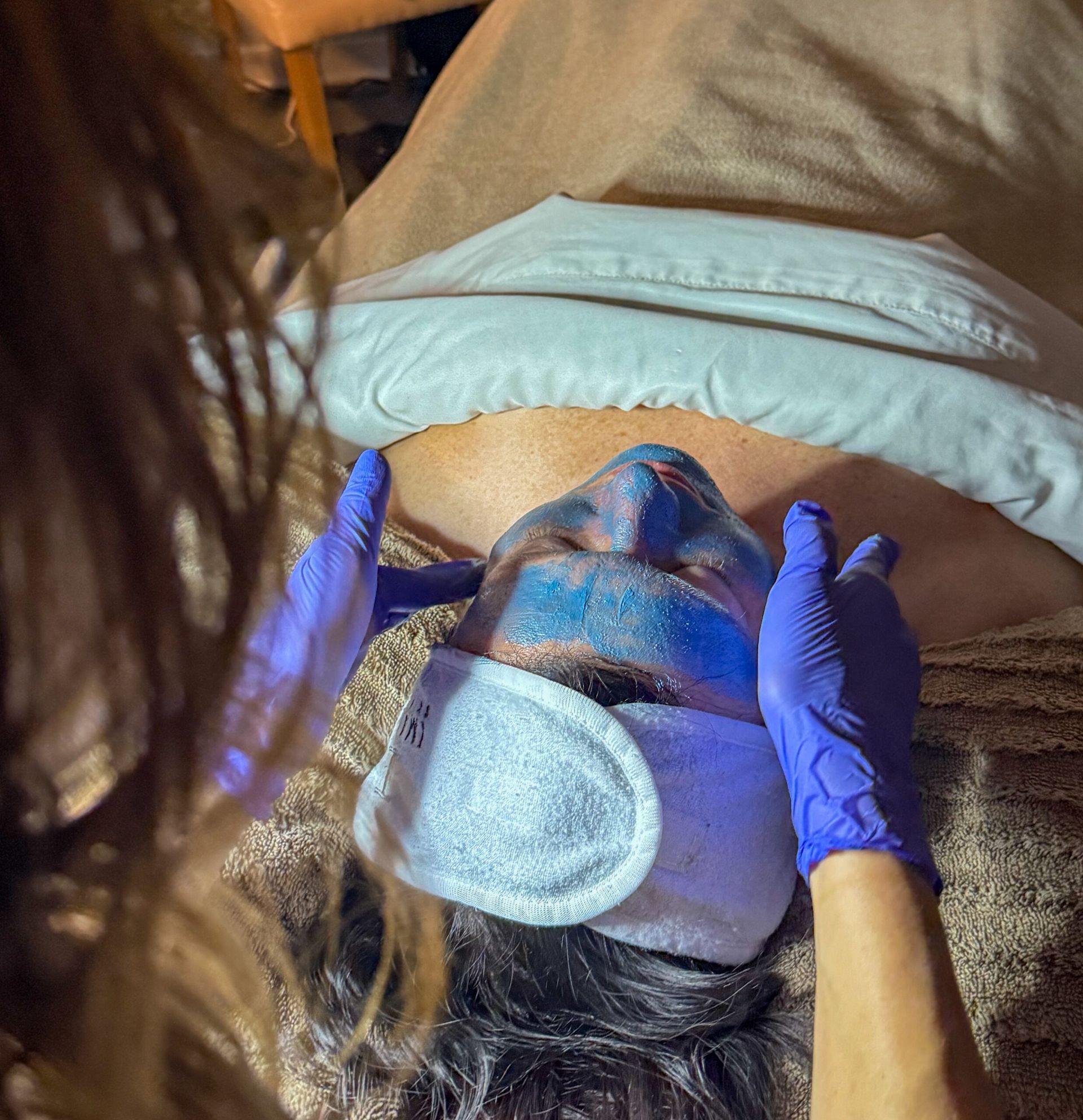 Person with blue face mask, receiving a spa facial, with gloved hands.