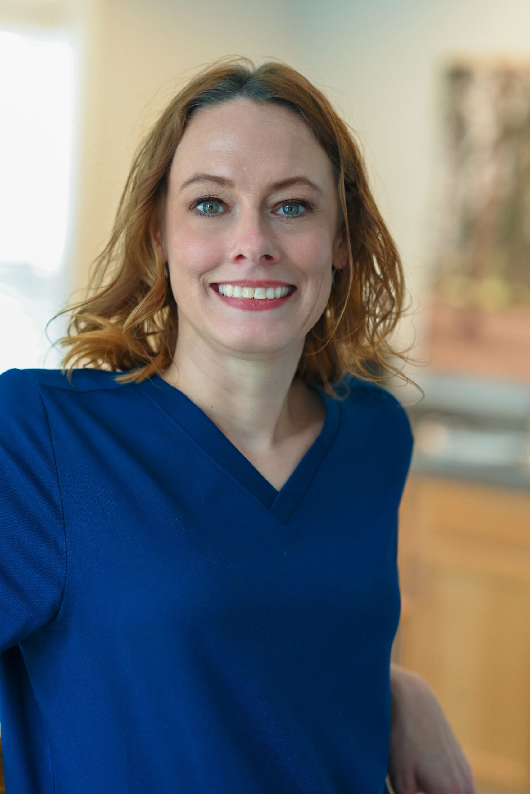 Woman with red hair smiles, wearing a blue sweater, indoors.
