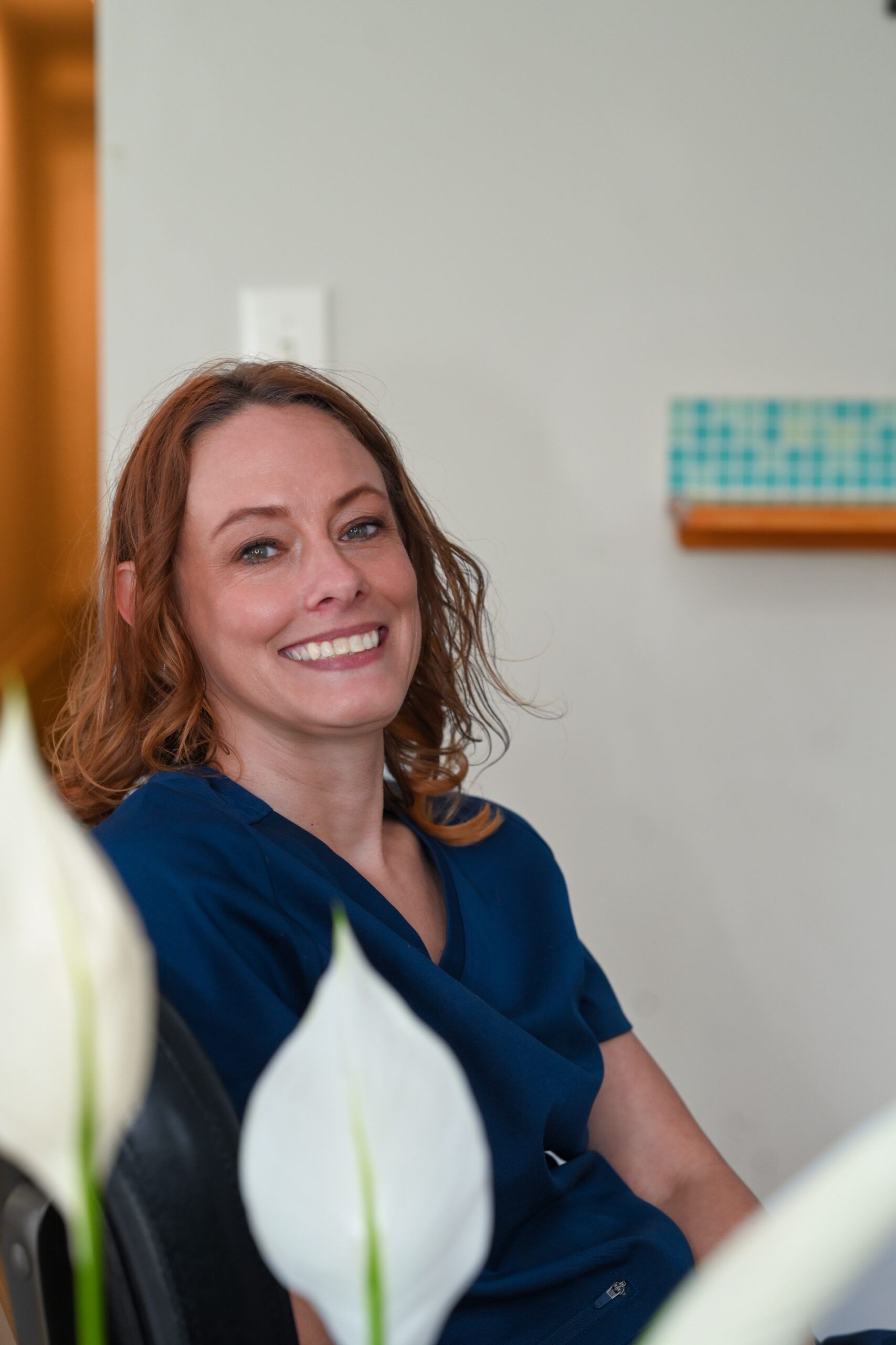 Woman with red hair smiles, wearing blue scrubs. Indoors, light background, white flowers in foreground.
