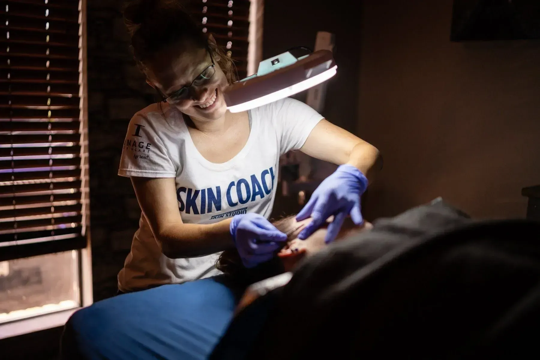 Woman, wearing gloves, performing skincare treatment under a magnifying lamp.