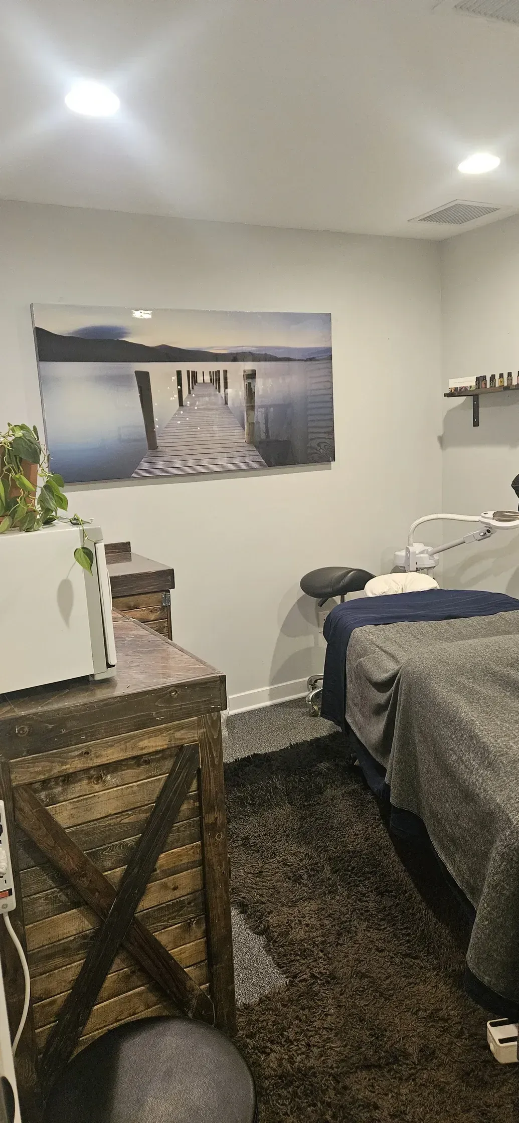 A massage room featuring a massage table with a grey blanket, wooden chest, and lake art on the wall.
