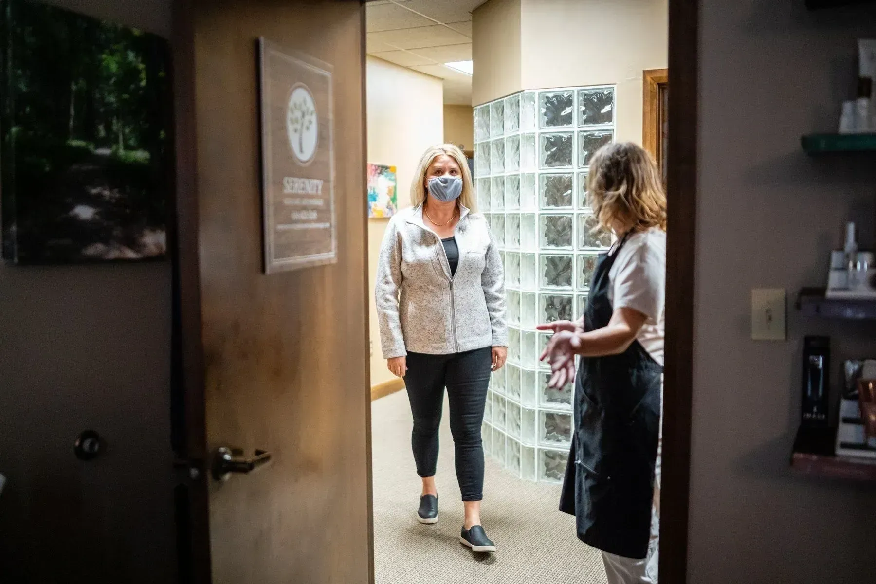 Woman in mask walks into a room; another woman in apron gestures. Interior office setting.
