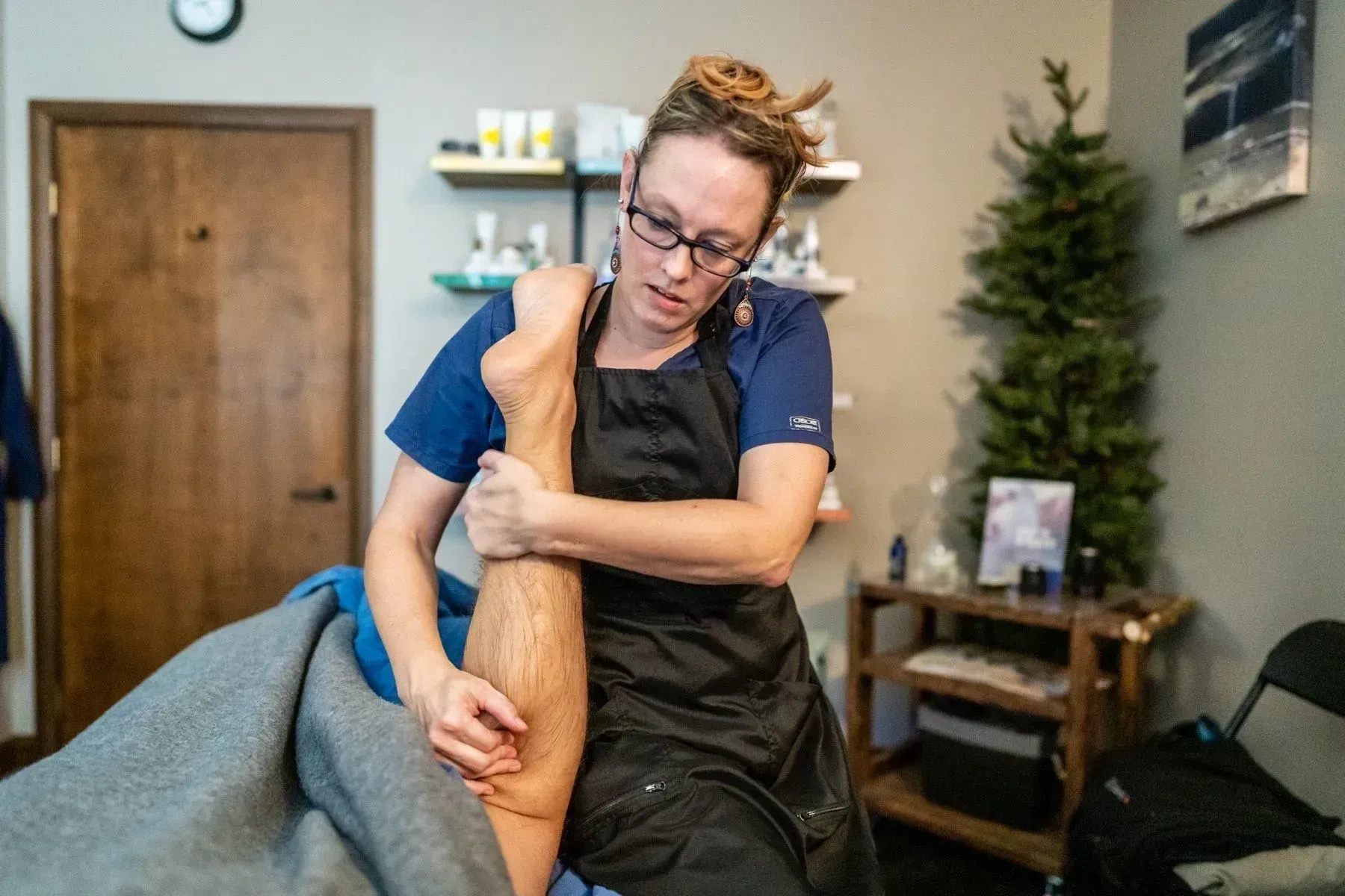 Massage therapist working on a patient's leg in a treatment room. The therapist wears glasses and a black apron.