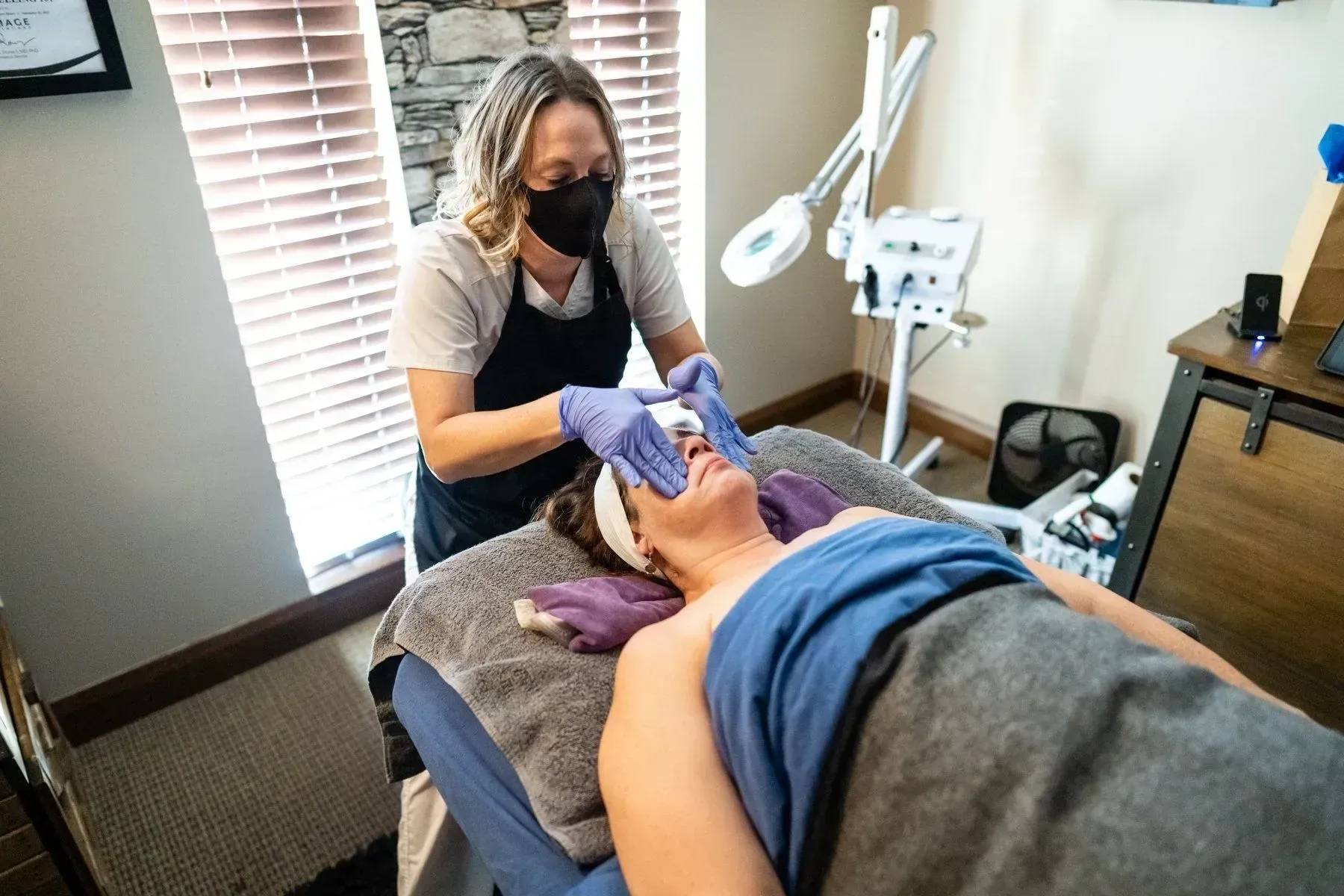 A masked esthetician performs a facial on a client, indoors.